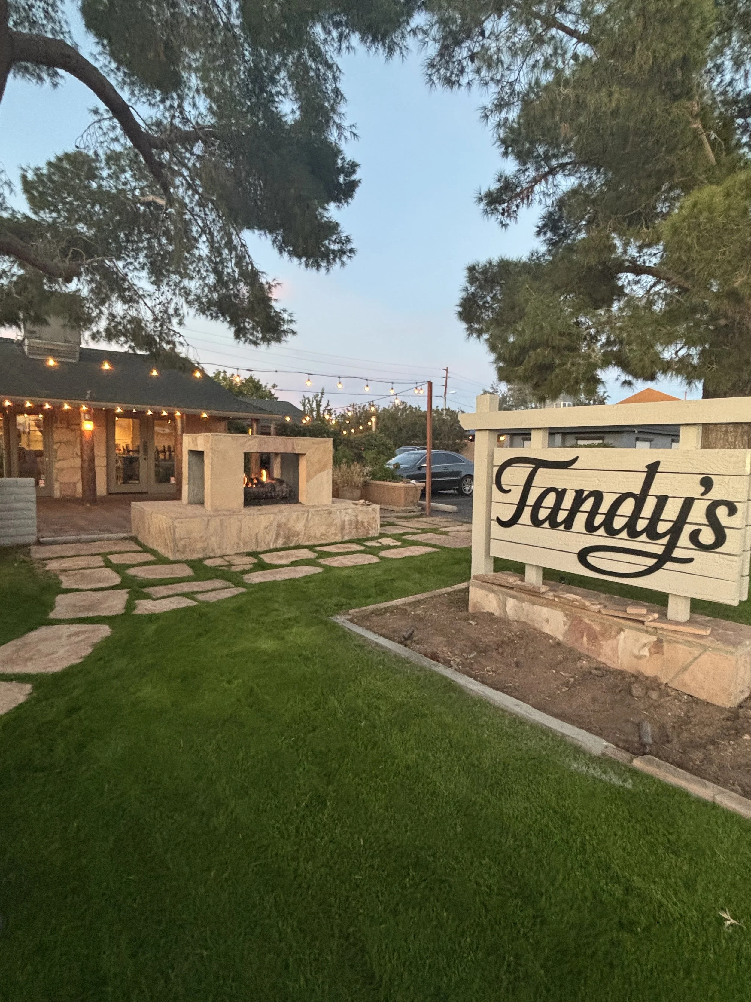 Outdoor scene at dusk with a large sign reading "Tandy's" in front of a building with string lights, a stone fireplace, and a grassy area with stone pathway.