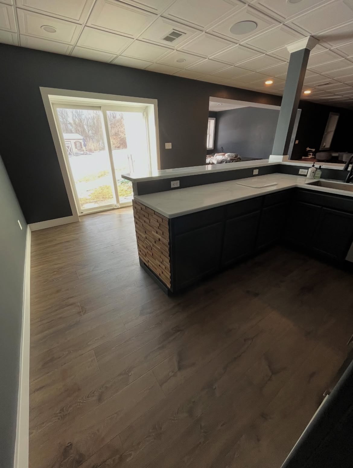 Interior view of a modern kitchen with dark cabinets, a white countertop, a brick accent, and a sliding glass door leading outside. There is a window in the background and wood-like flooring.