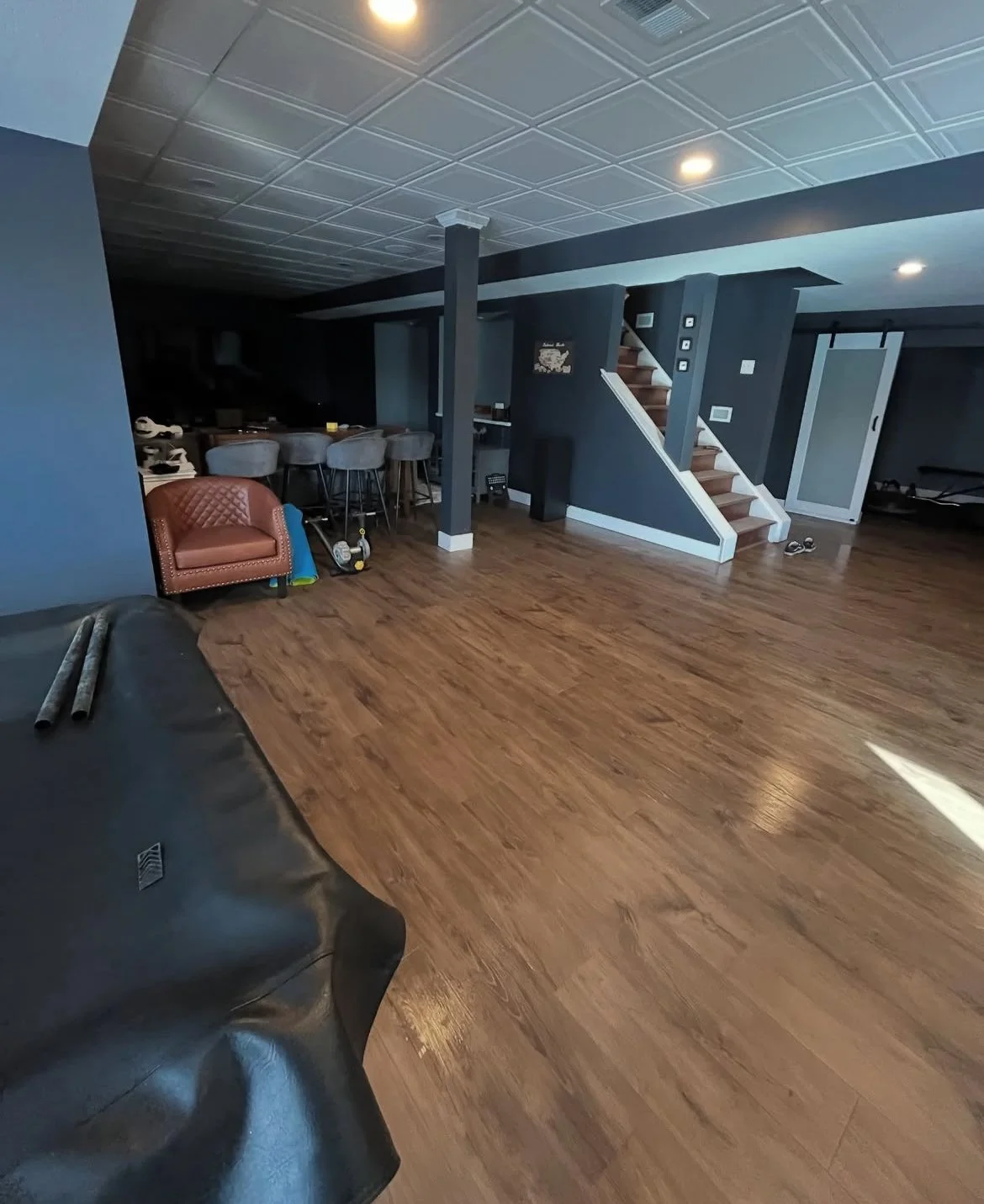 Interior view of a basement with dark blue walls, a staircase, a bar area with bar stools, and hardwood flooring.