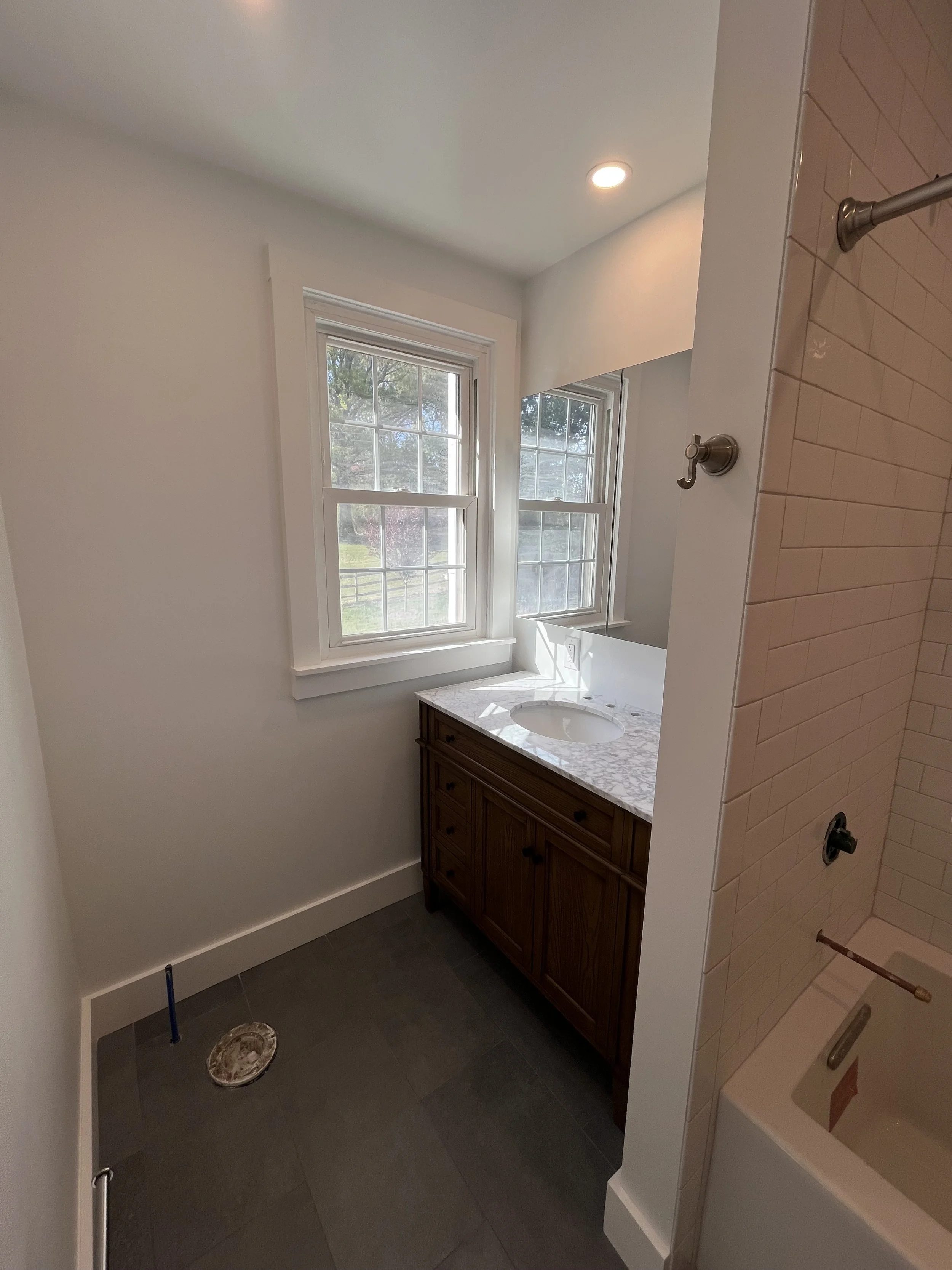 New bathroom with two windows, a wood vanity with marble countertop and sink, a wall mirror, and a separate shower area with beige subway tile, showerhead, and frosted glass door.