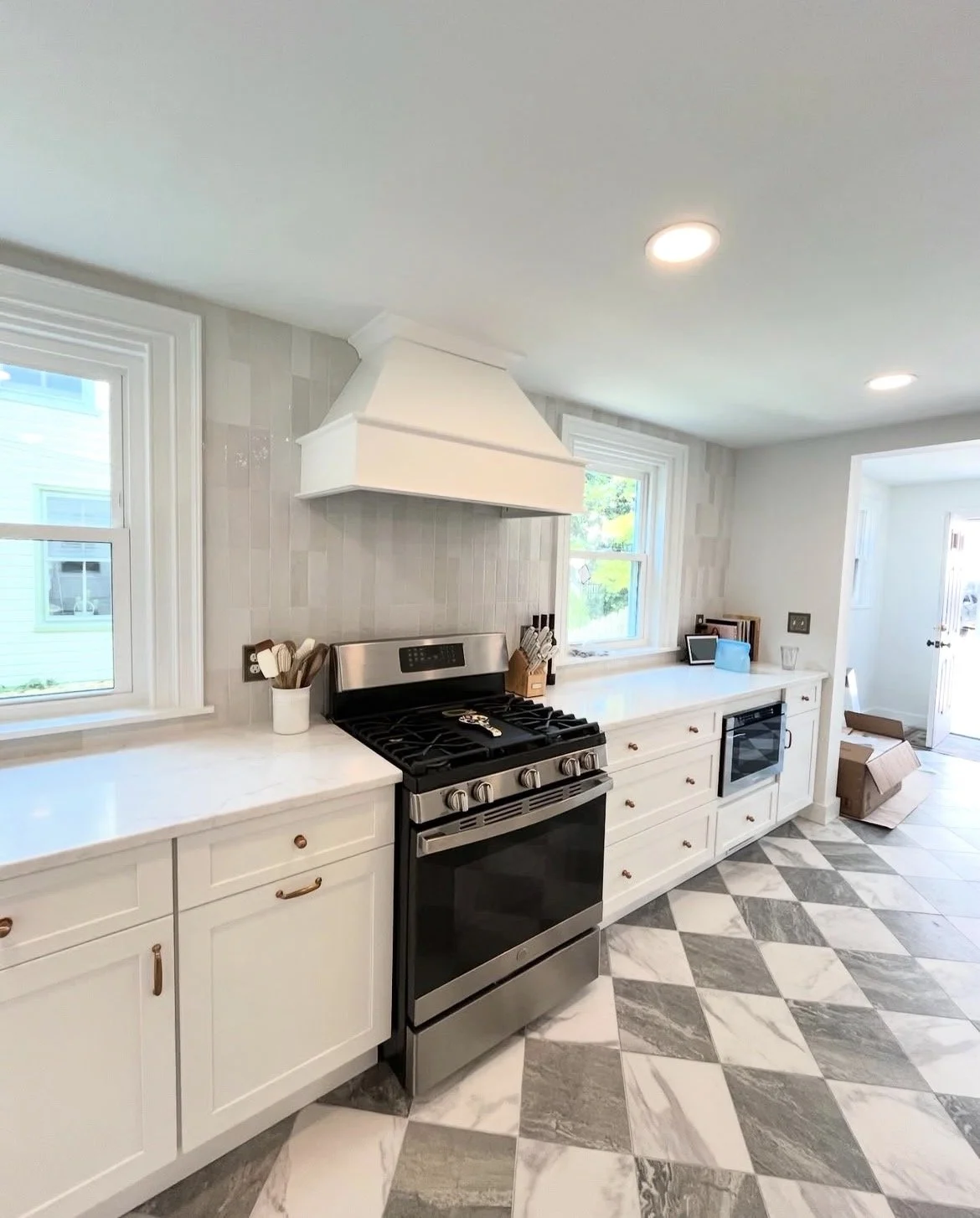 Modern kitchen with white cabinets, stainless steel oven, black cooktop, marble patterned floor, windows, and minimalist decor.