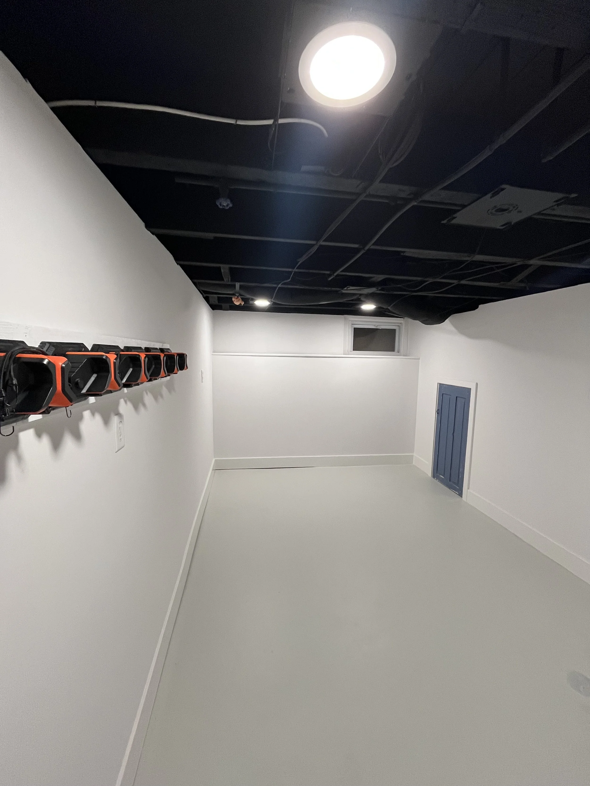 Empty room with white walls, a row of orange and black tools on the left wall, small window near the ceiling, and a blue door on the right wall. The ceiling is unfinished with visible wires and lighting.