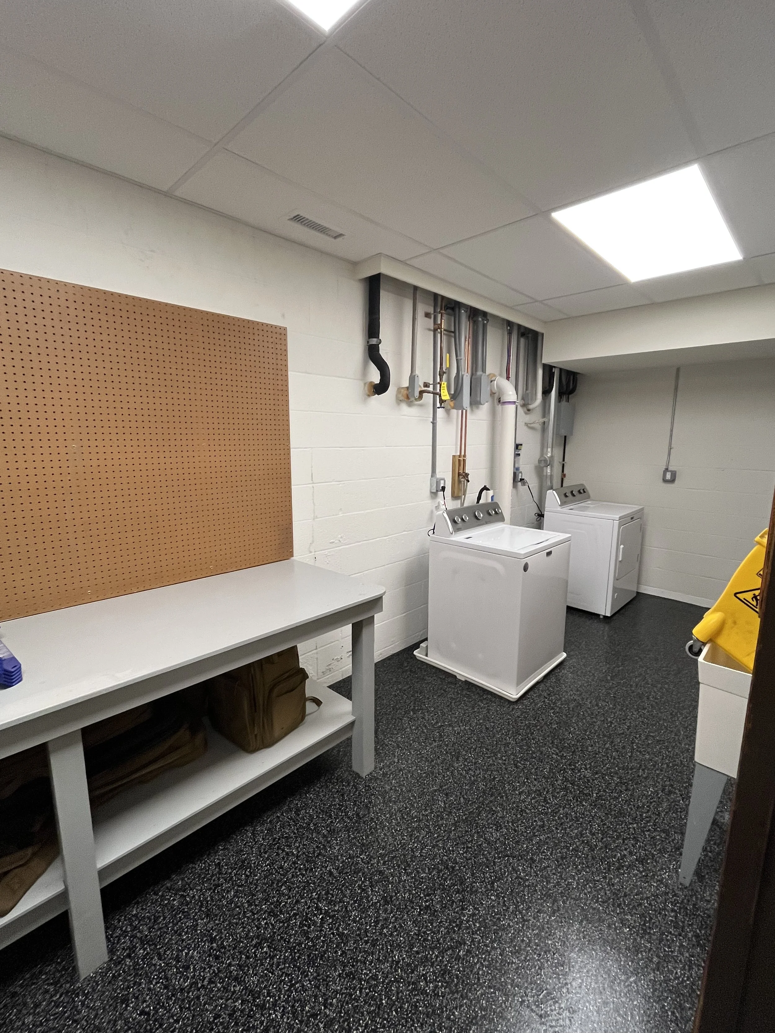 A workroom with a speckled black floor, white walls, and a white ceiling with fluorescent lights. There are washers and dryers in the corner, a pegboard on the wall, and a white table with brown bags underneath.