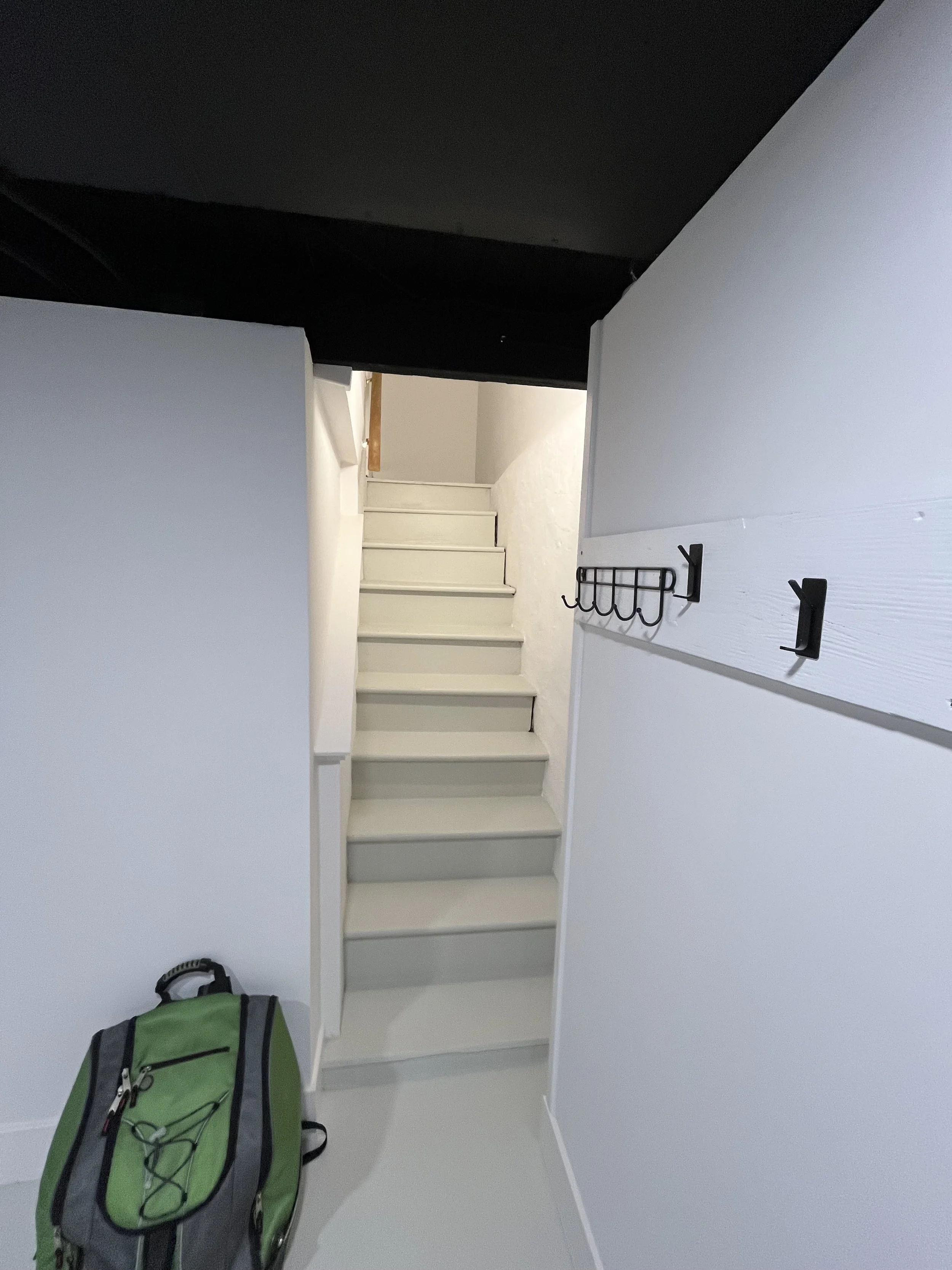 A small white staircase with six steps, white walls, a black hook rack on the right wall, and a green and gray backpack on the floor near the stairs. The ceiling is black and the floor is white.
