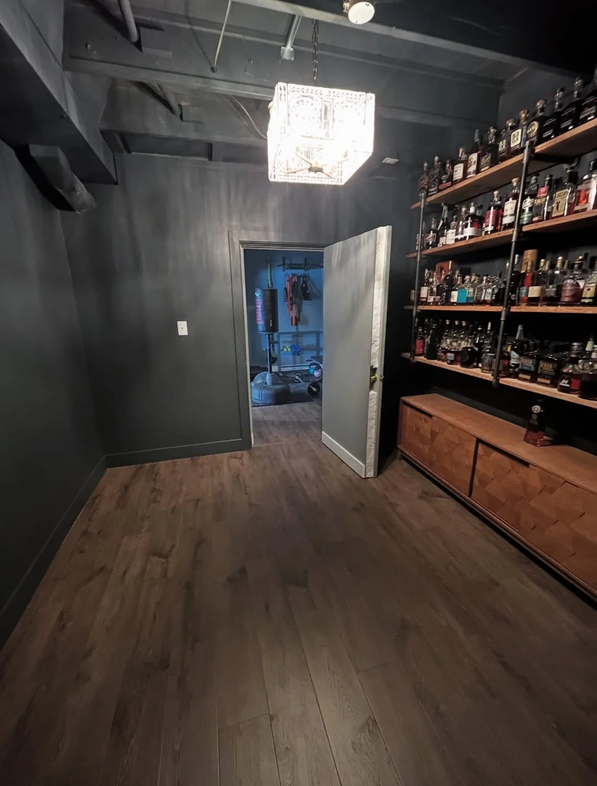 A room with dark gray walls and wooden flooring, featuring a ceiling light fixture, a doorway revealing a storage room with gym equipment, and a wooden shelving unit filled with liquor bottles. Expert design and remodel by MGD Carpentry