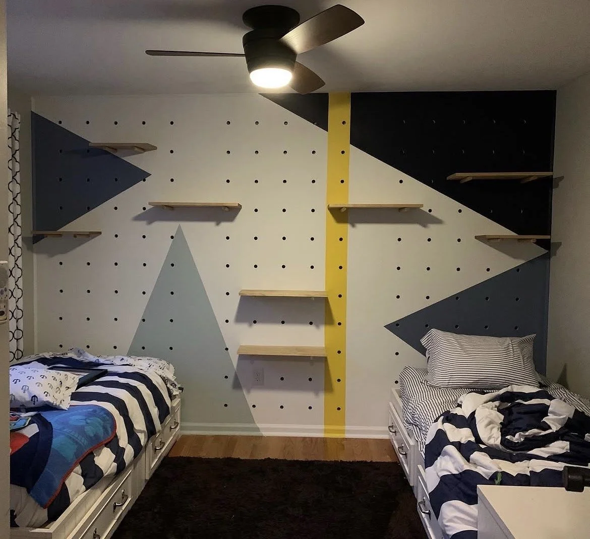 Children's bedroom with a geometric painted wall, three beds with striped bedding, and wall-mounted shelves.