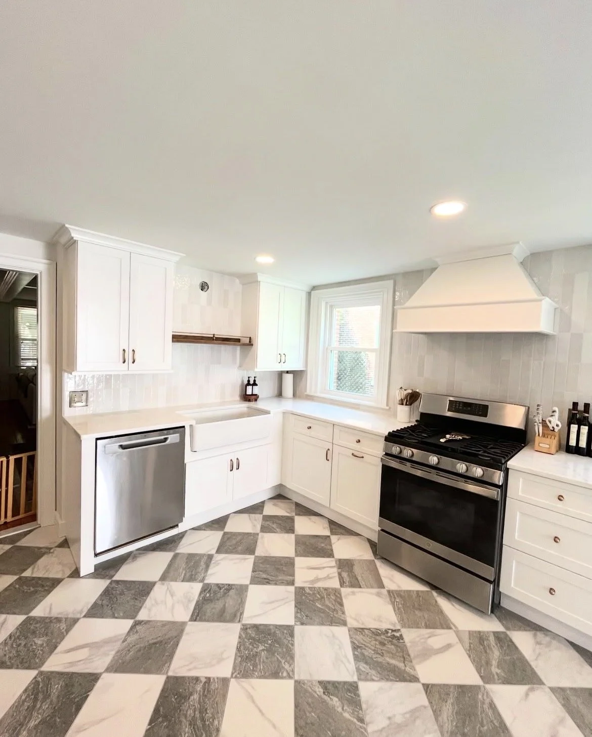 Modern white kitchen with black and white checkered marble floor, white cabinets, stainless steel appliances, and a window above the countertops.