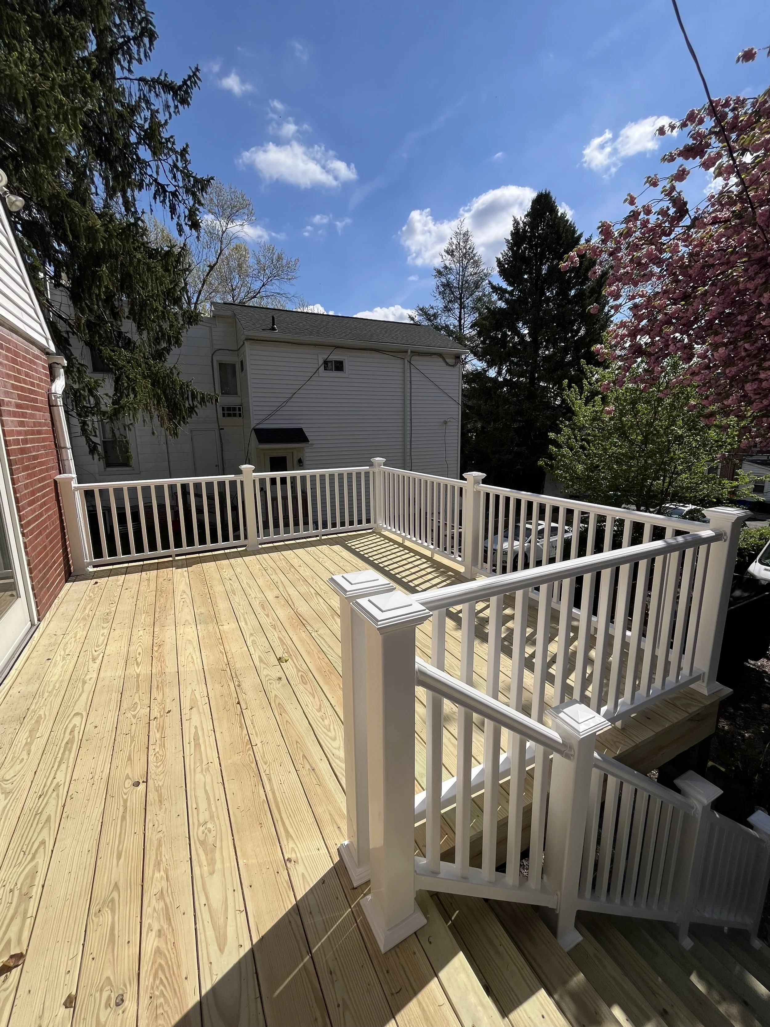 A newly built wooden deck with white railing surrounds the back of a house, overlooking neighboring houses and trees, with a bright blue sky and some clouds overhead.