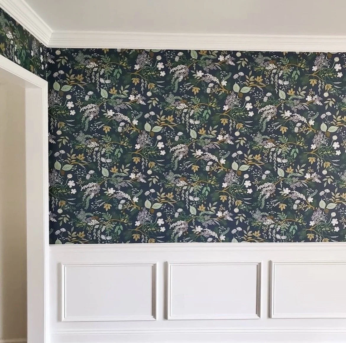 Close-up of a wall with dark floral wallpaper featuring white, purple, and gold flowers and green leaves, with white wainscoting below.