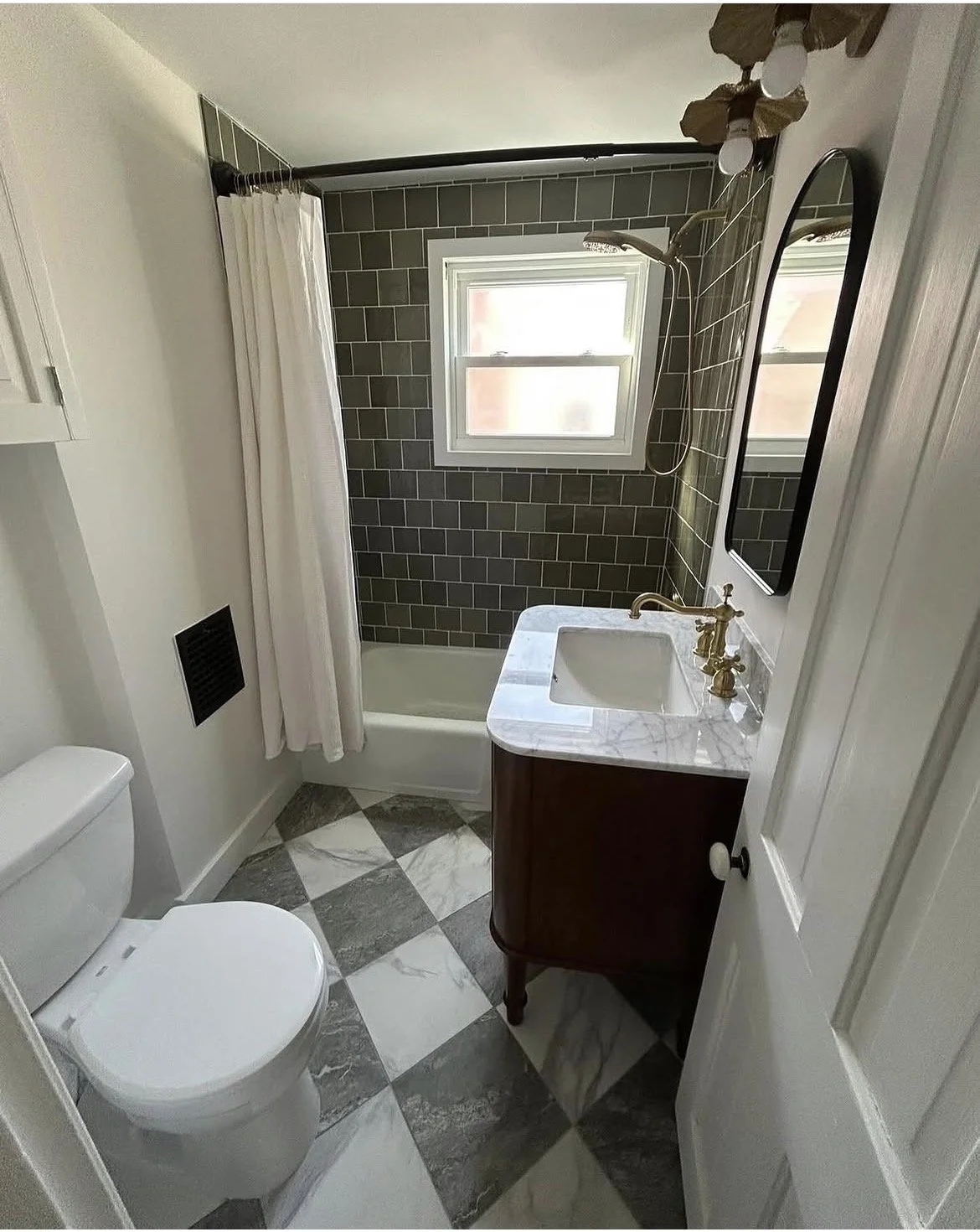 A small bathroom with a toilet, a vanity with a marble top and brass faucet, a bathtub with a green tiled wall, a window, and a mirror.