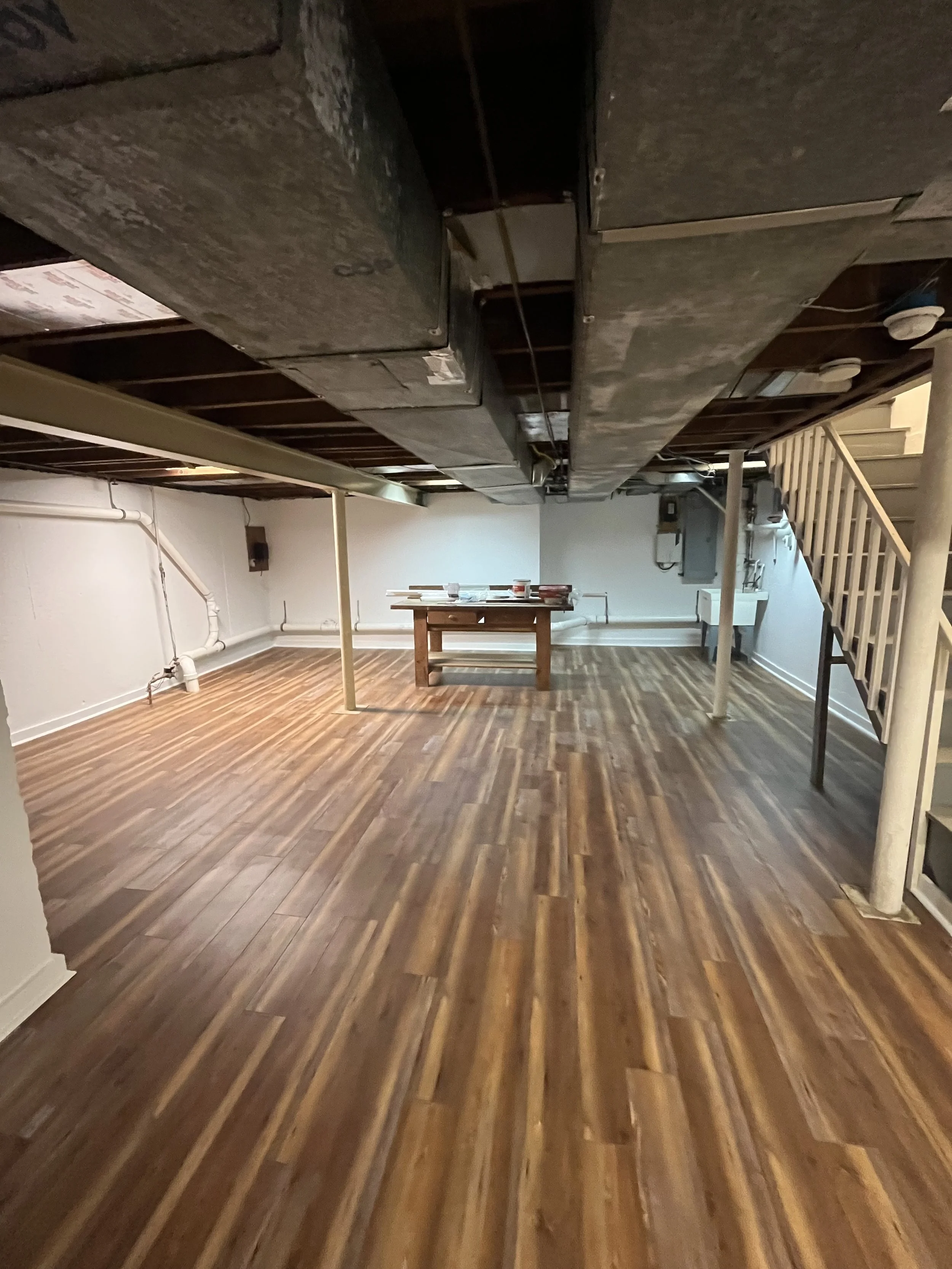 Basement room with unfinished ceiling, wood staircase, hardwood floor, and white walls, featuring visible pipes and electrical wiring.