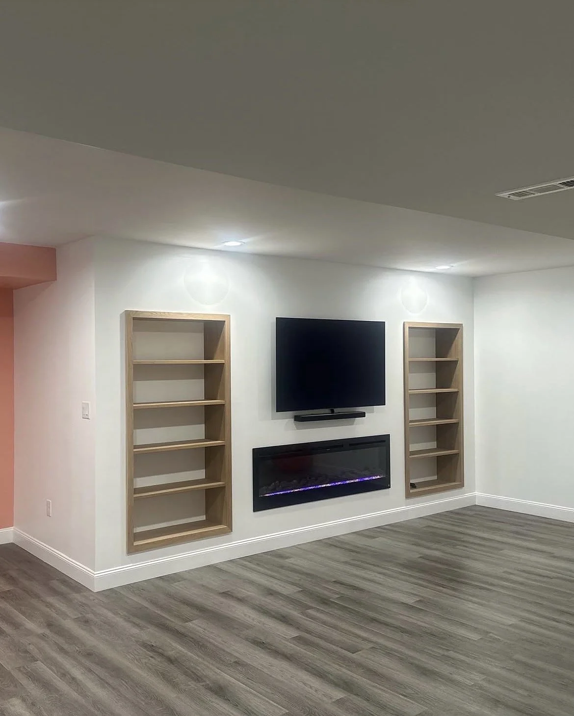 Living room corner with wall-mounted TV, electric fireplace, and two empty wooden bookshelves on either side, with hardwood flooring and white walls.