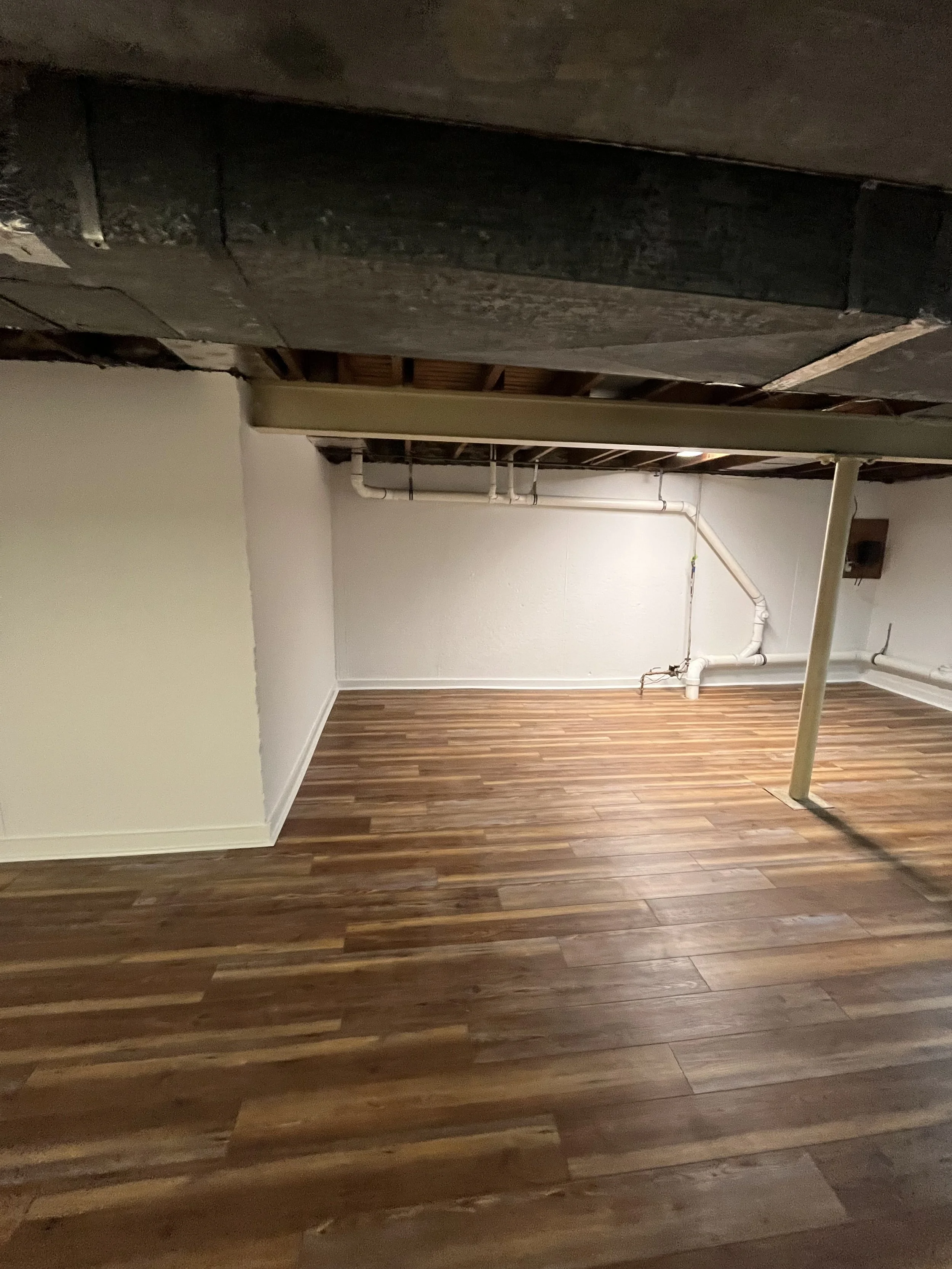 A basement or unfinished room with hardwood flooring, white and beige walls, and exposed ceiling beams and pipes.