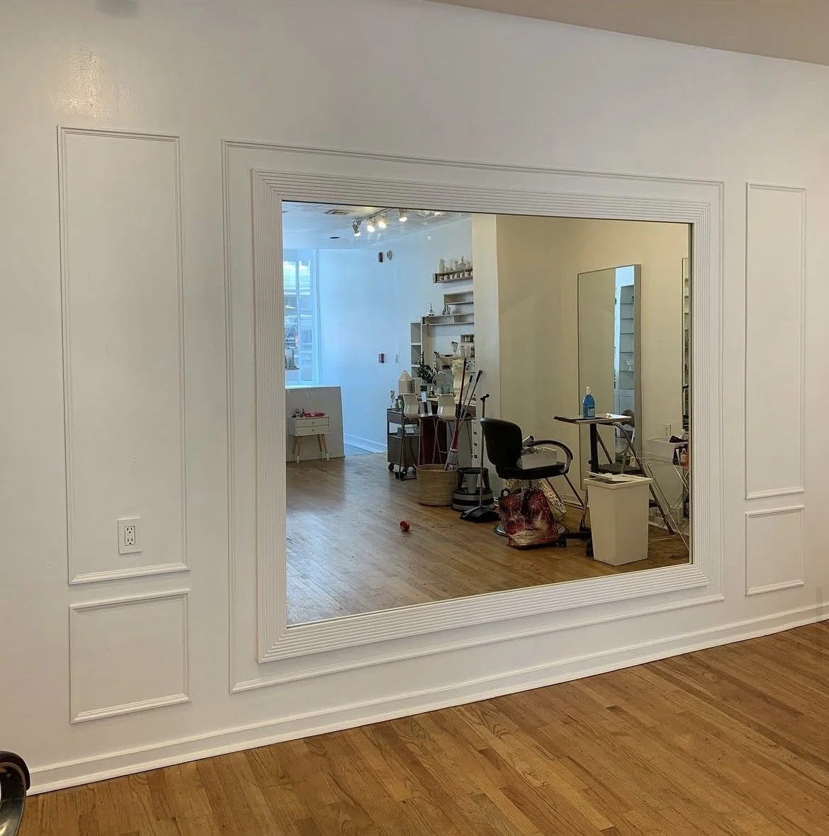 Empty room with a large framed mirror on a white wall, reflecting a cluttered space with furniture, cleaning supplies, and a window in the background.