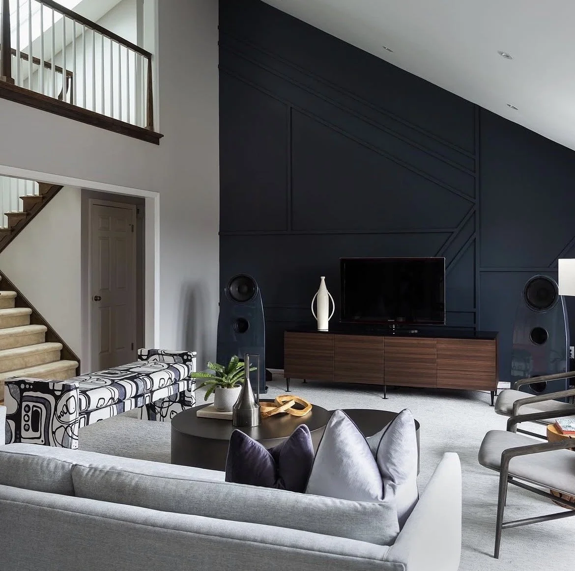 Modern living room with gray sofa, black and white patterned bench, coffee table with decor, flat-screen TV on wooden cabinet, dark blue accent wall, two large speakers, staircase with beige carpet, and minimal decor.