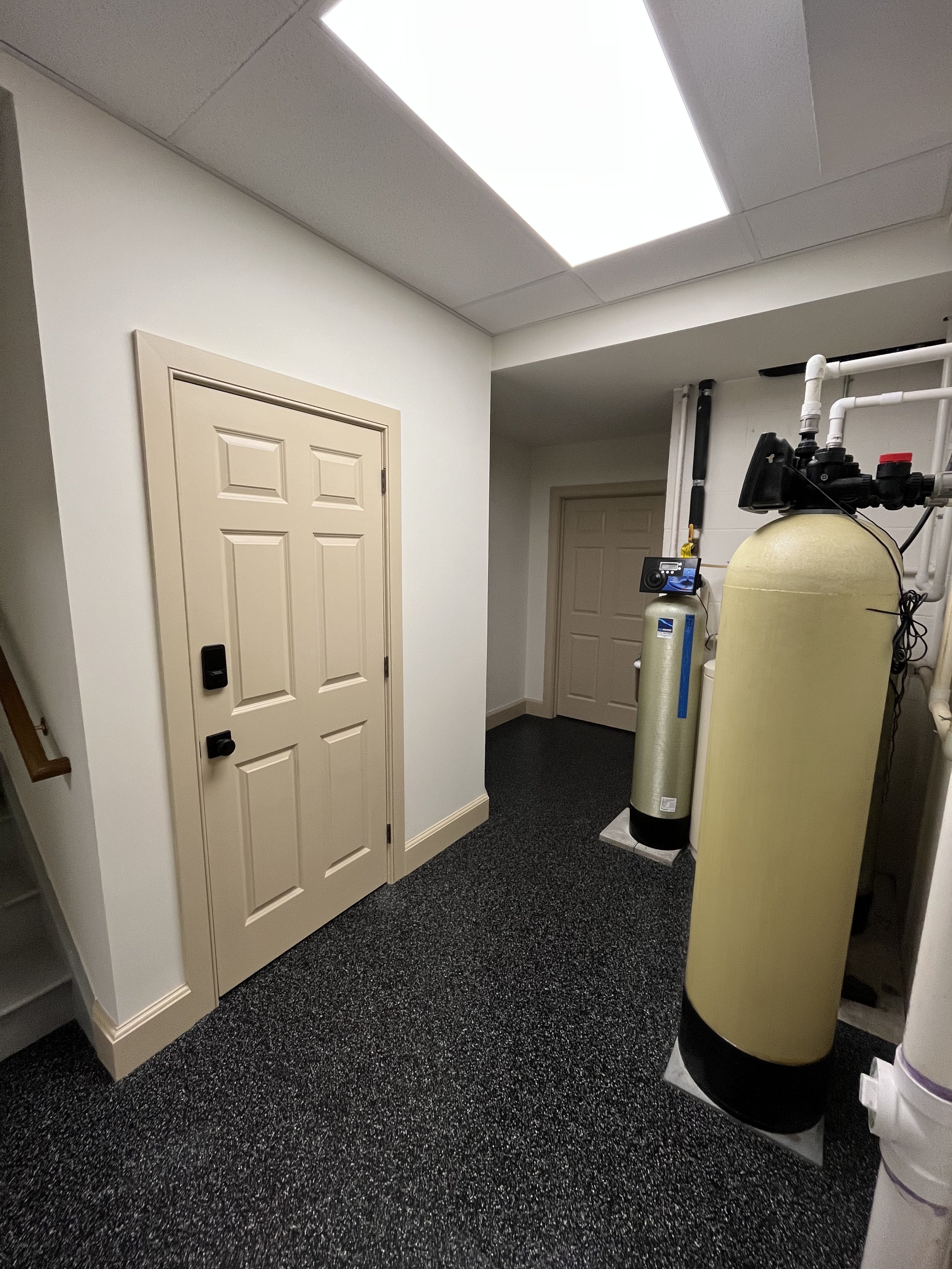 Interior of a utility or mechanical room with water filtration or water softening system, including large cylindrical tanks, pipes, and a control panel, with beige walls and dark speckled carpeting.