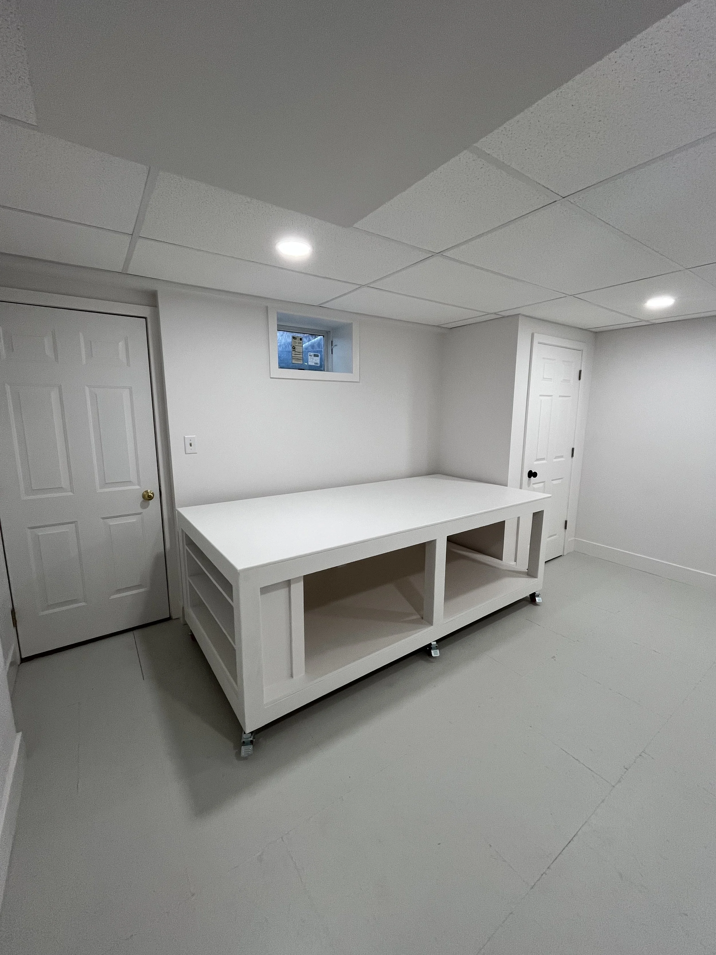 A white table on casters in a basement room with white walls, a small window, and a closed white door.
