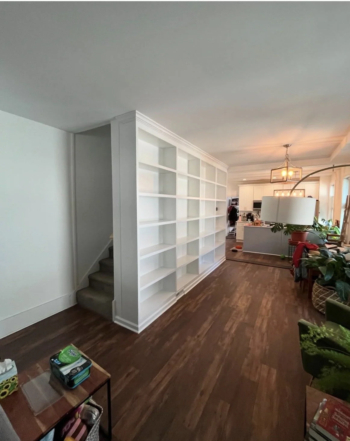 Interior view of a house with hardwood floors, a white built-in bookshelf dividing the living room and kitchen, a staircase on the left, and various furniture and plants in the living area.