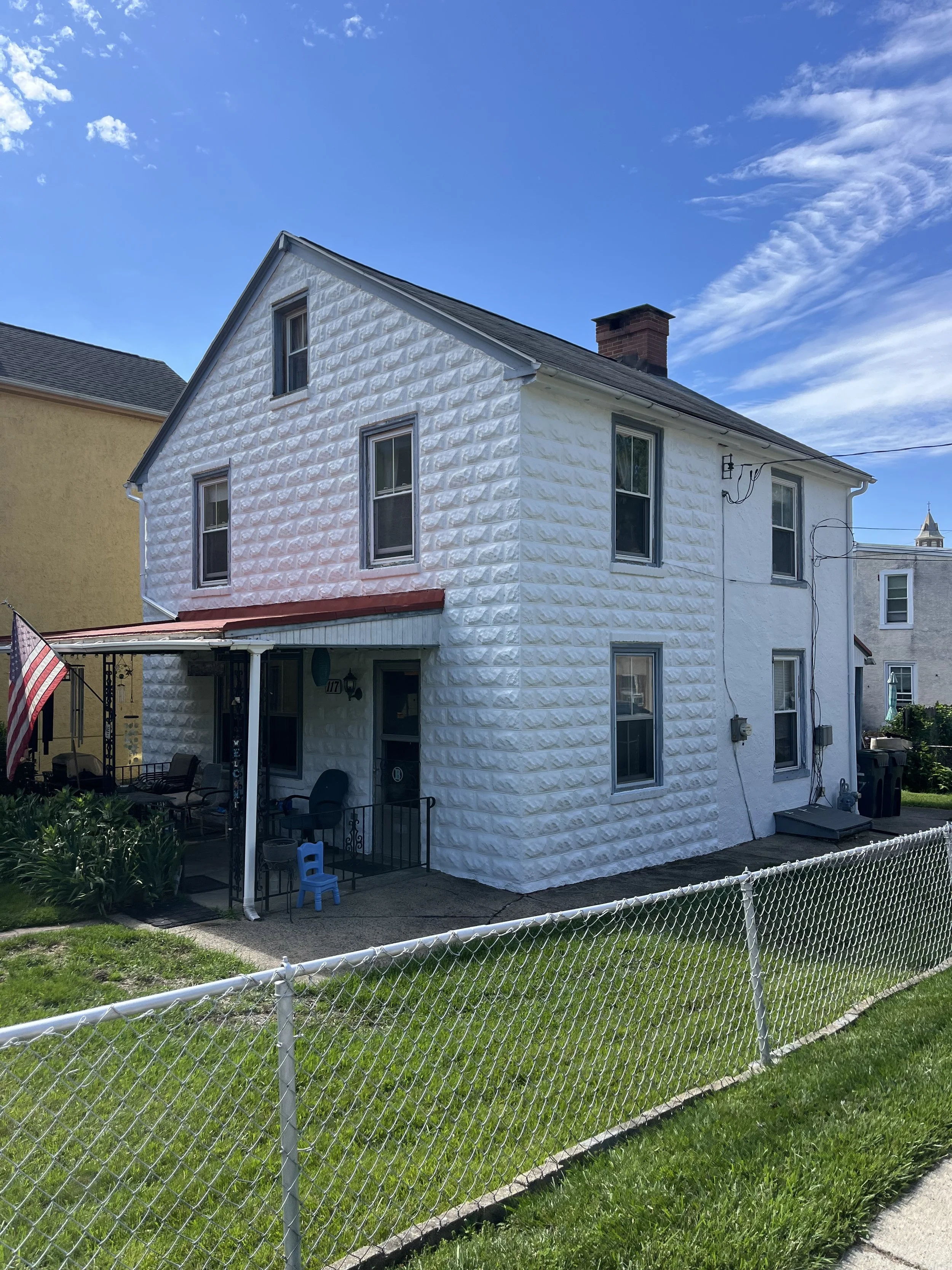 A white, two-story house with a sloped roof, multiple windows, a small front yard enclosed by a white picket fence, and a backyard with patio furniture and an American flag, under a partly cloudy blue sky.