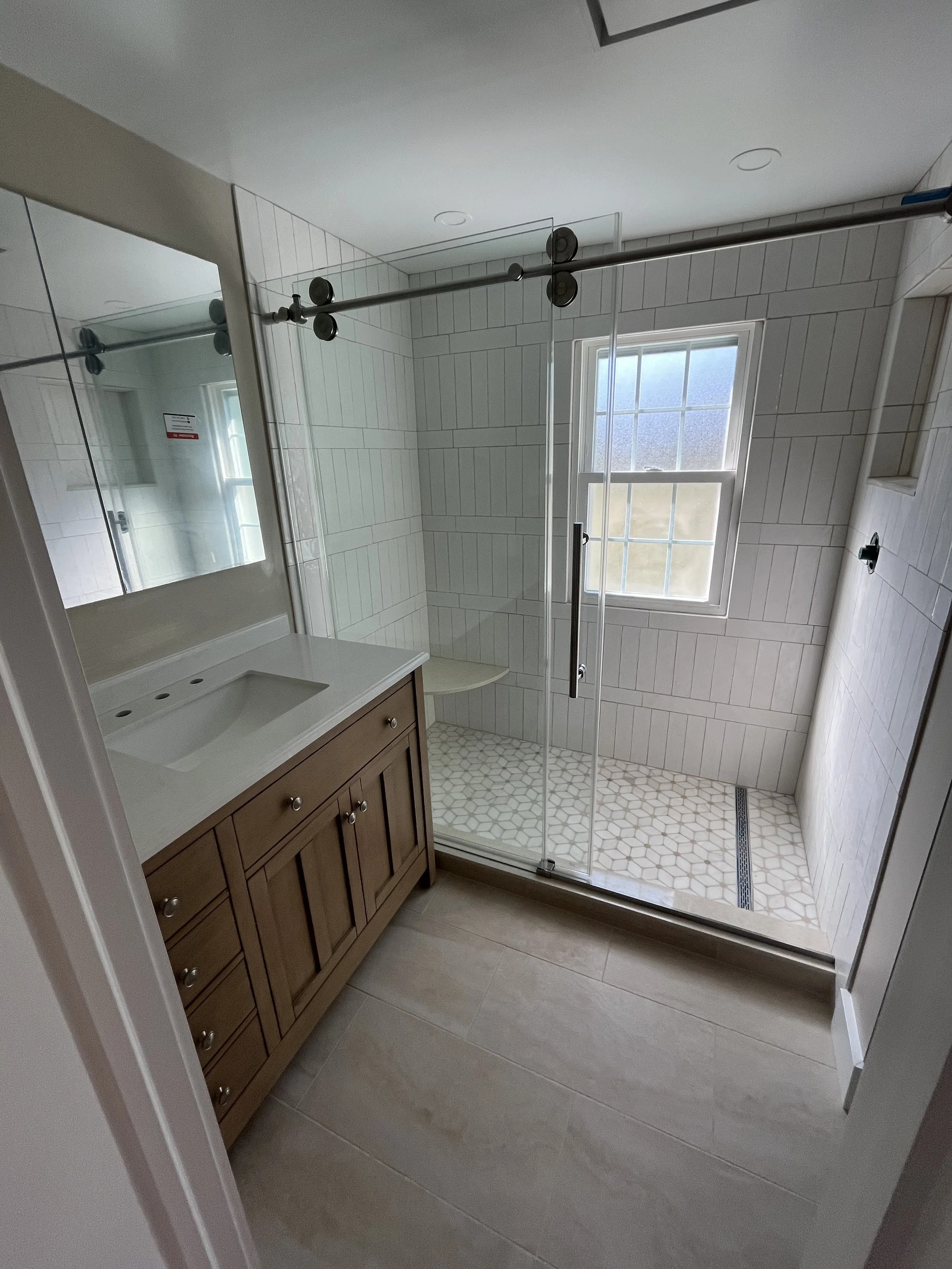 Modern bathroom with a glass-enclosed shower, a window, a wooden vanity with a white sink, and beige floor tiles.