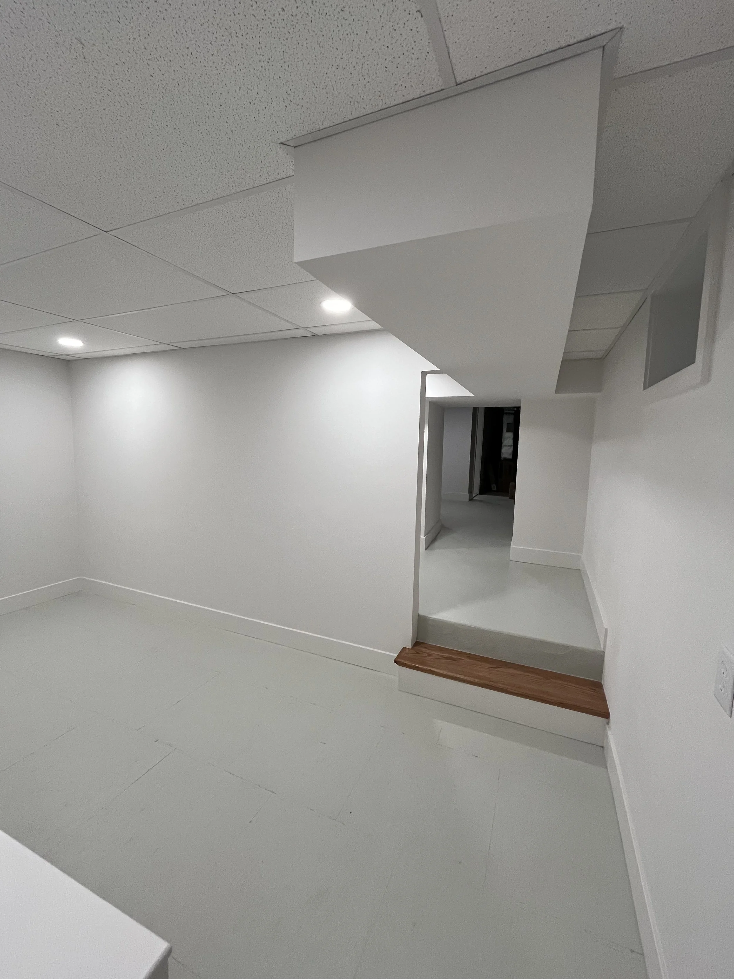 Modern, empty room with white walls, suspended ceiling tiles, and ceiling lights. A small wooden platform or step is visible near the corner.