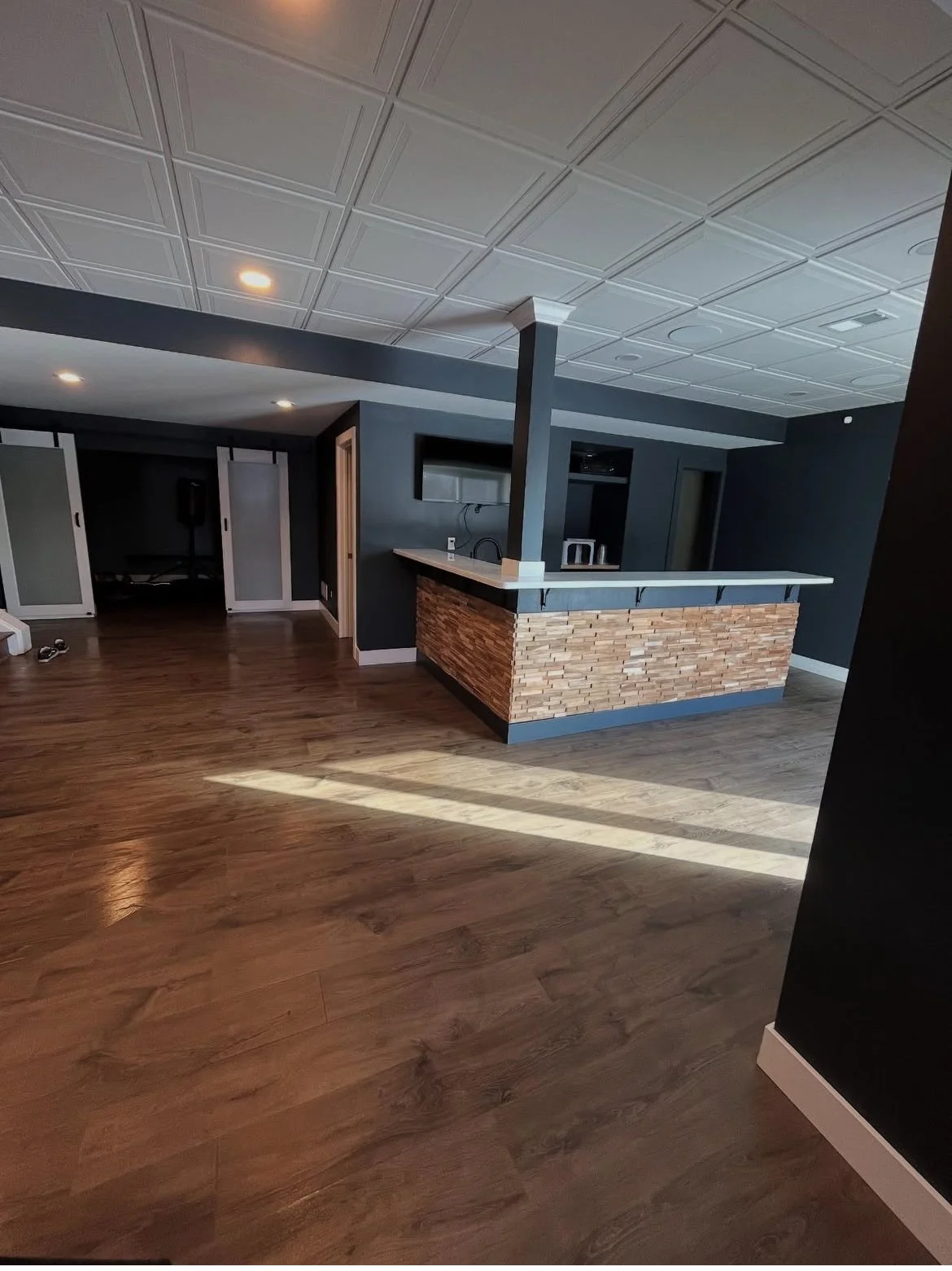 Interior view of a modern basement with dark walls, wooden flooring, a bar with brick facade, and a ceiling with white tiles. Sunlight streams in, casting shadows on the floor.