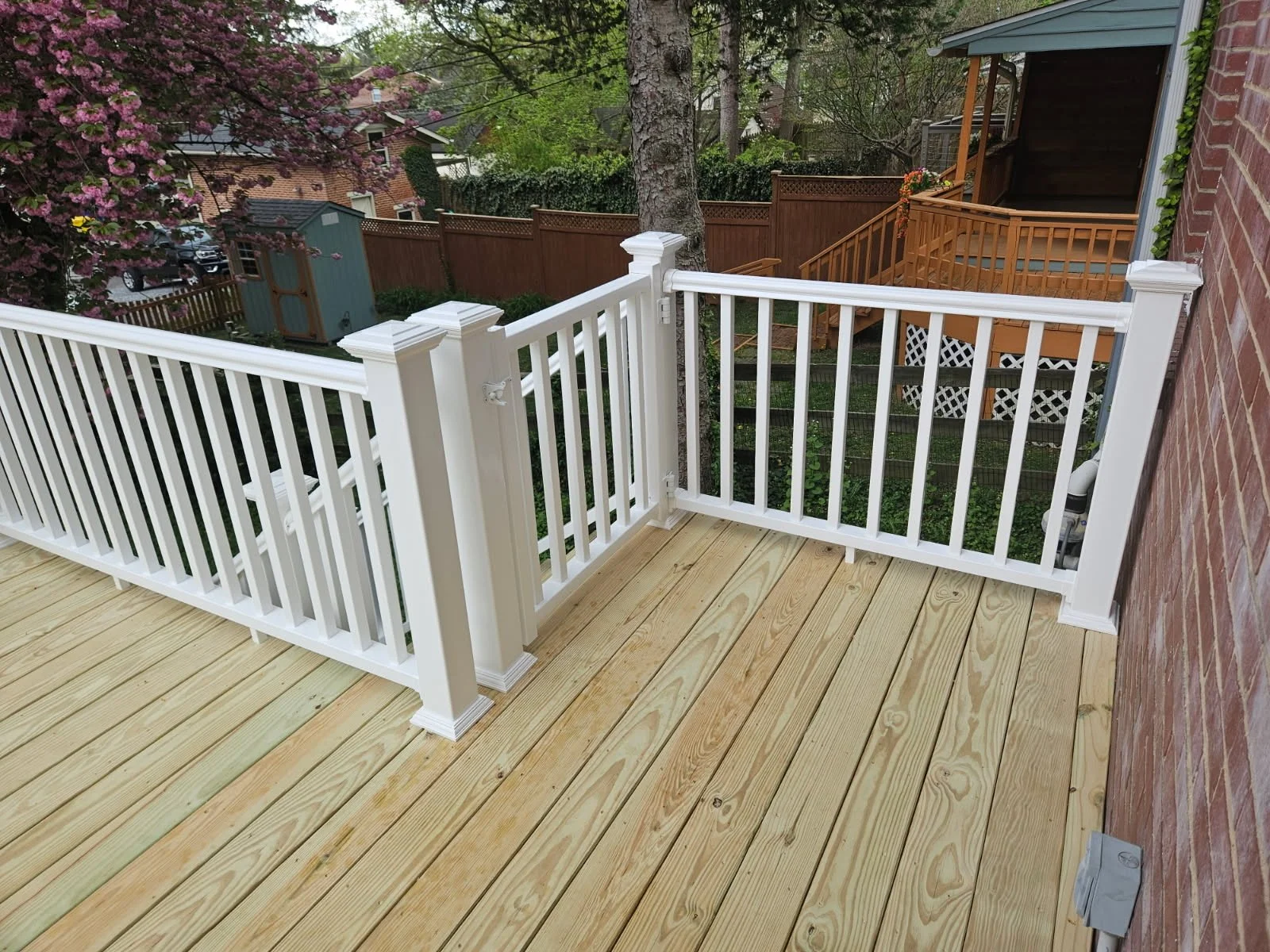 View from a porch with newly installed wooden floorboards and a white railing overlooking a backyard with trees, a fence, and a neighboring house.