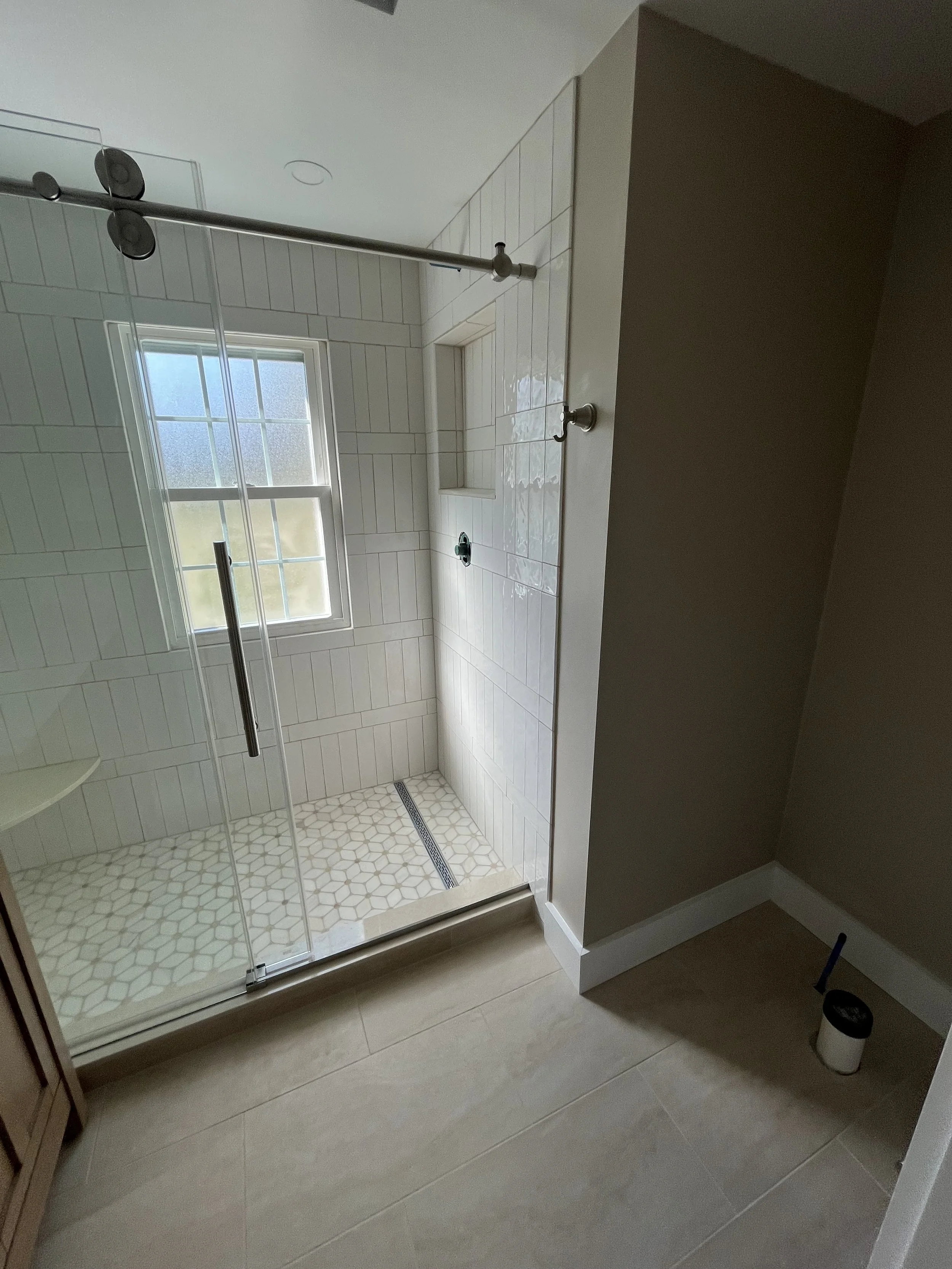 A bathroom shower with a clear glass door, white tile walls, and a small window. Inside the shower, there is a built-in niche, a black faucet, and a patterned hexagonal floor drain. Outside the shower, there is a beige floor with large tiles, a woode