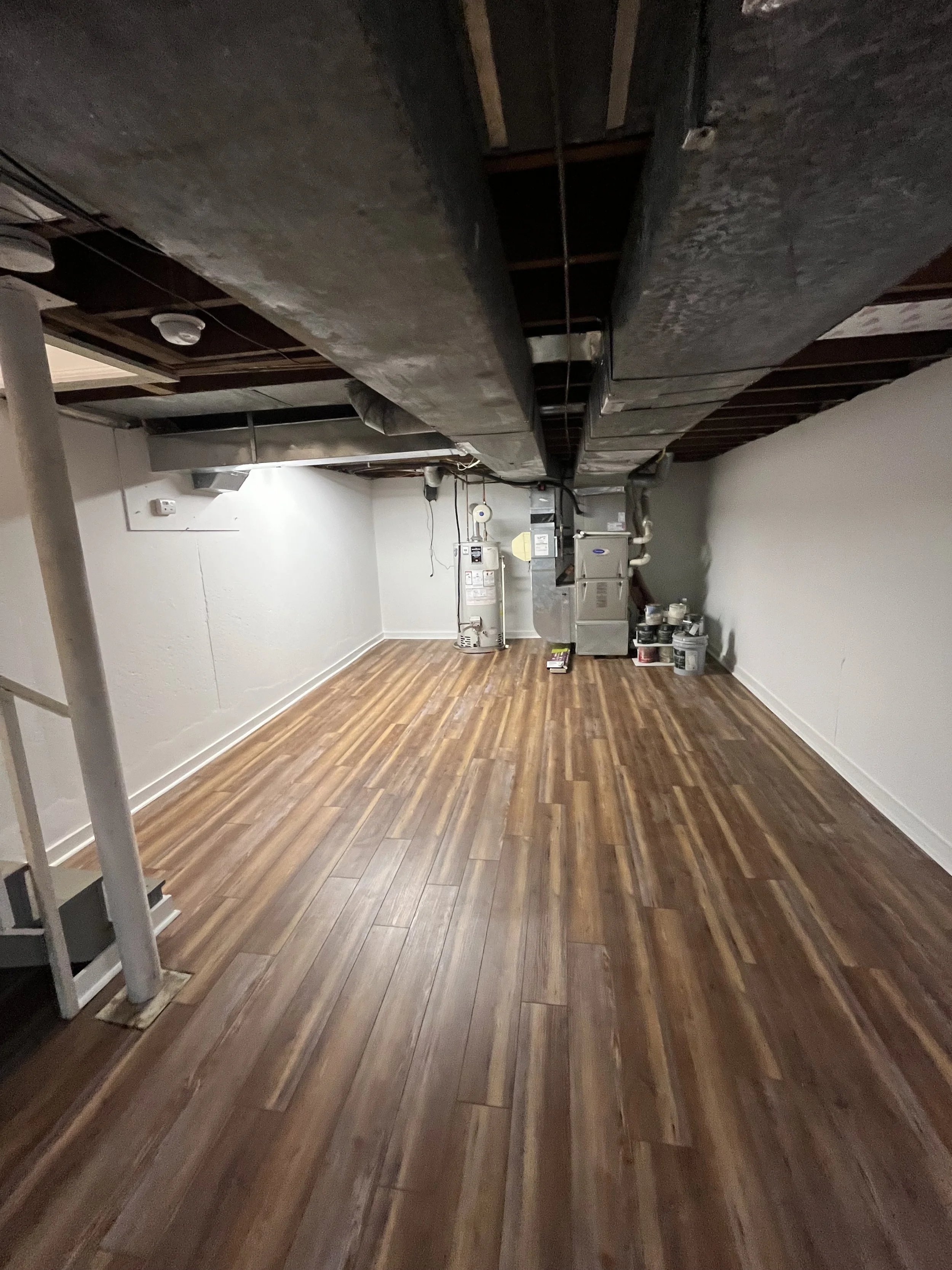 A room with brown wooden floor, white walls, exposed ceiling beams, HVAC system, and containers of paint or supplies in the corner.