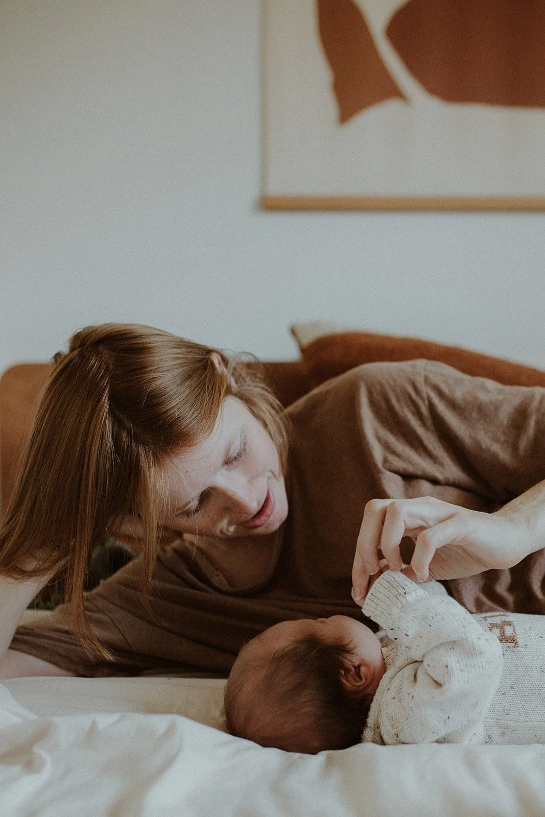 A young woman with red hair lying on a bed, looking at a sleeping baby. The woman appears to be gently touching the baby's hand. The scene is cozy, with warm lighting and a neutral-colored background.