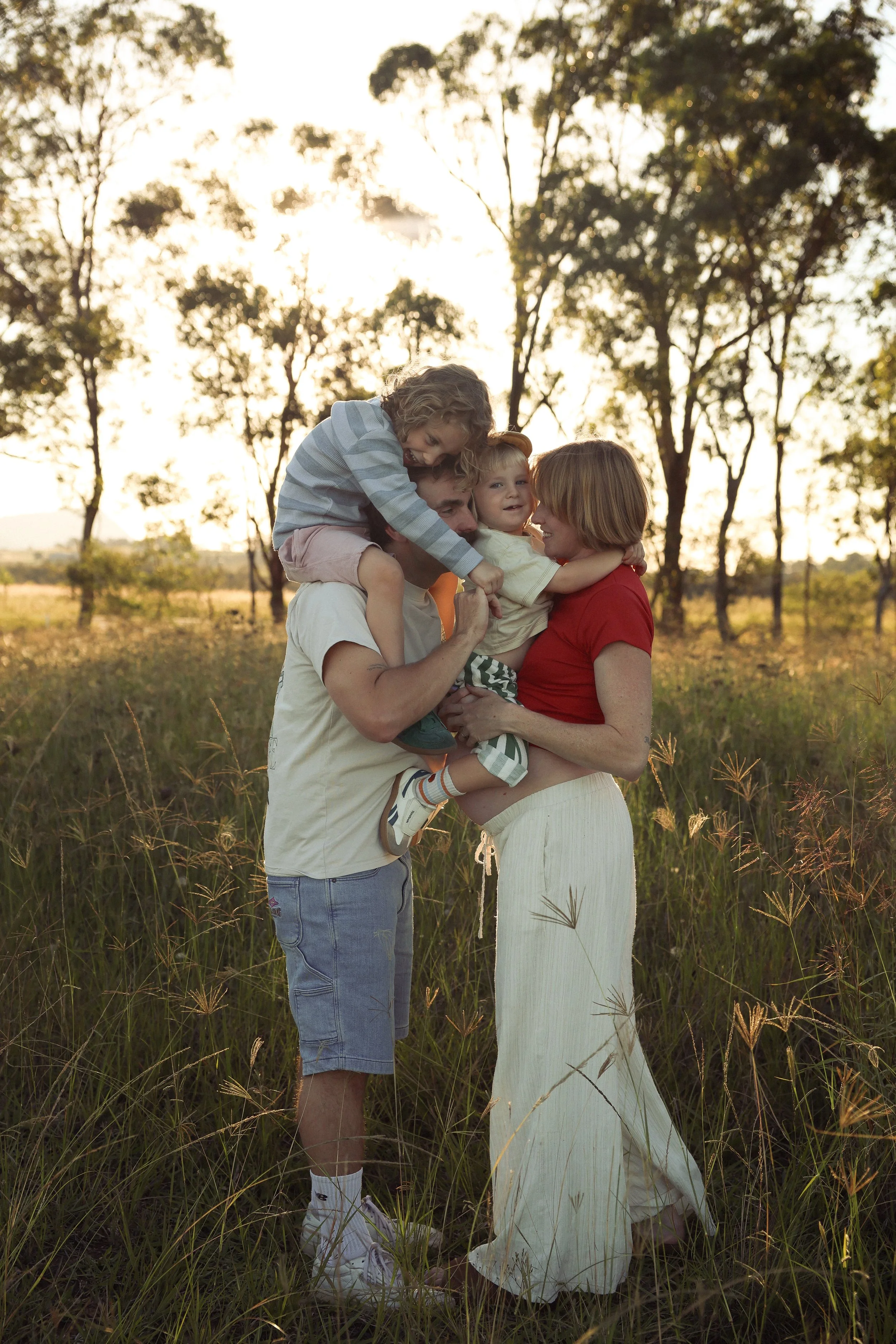 A family of four, including a man, woman, and two young children, enjoying a moment together outdoors in a grassy field with trees and sunset in the background.