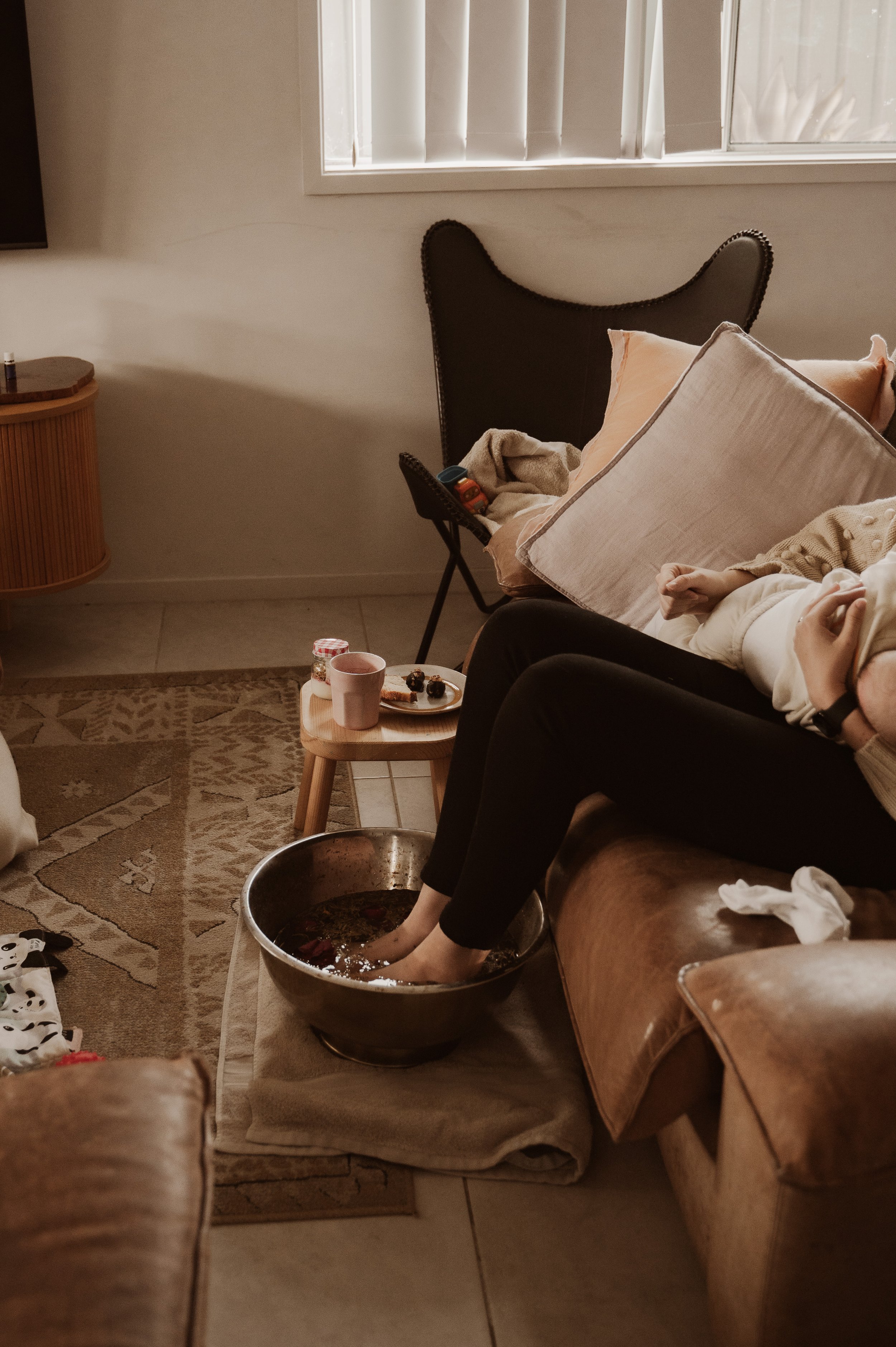 Person soaking feet in a foot bath on a living room floor, with pillows on their lap, a table with a mug and food nearby, and a window in the background.