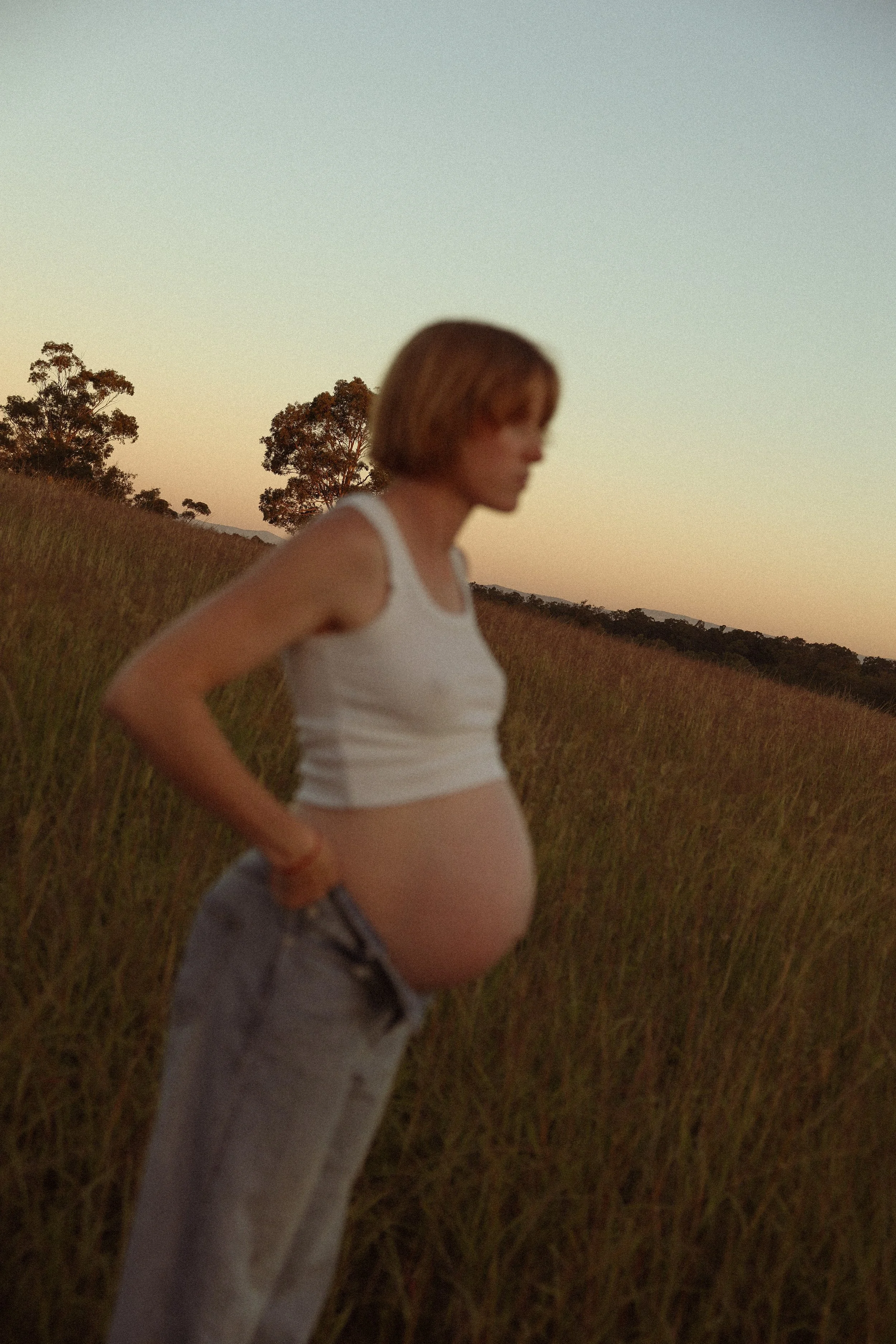 A pregnant woman standing in a field during sunset, wearing a white tank top and gray pants.