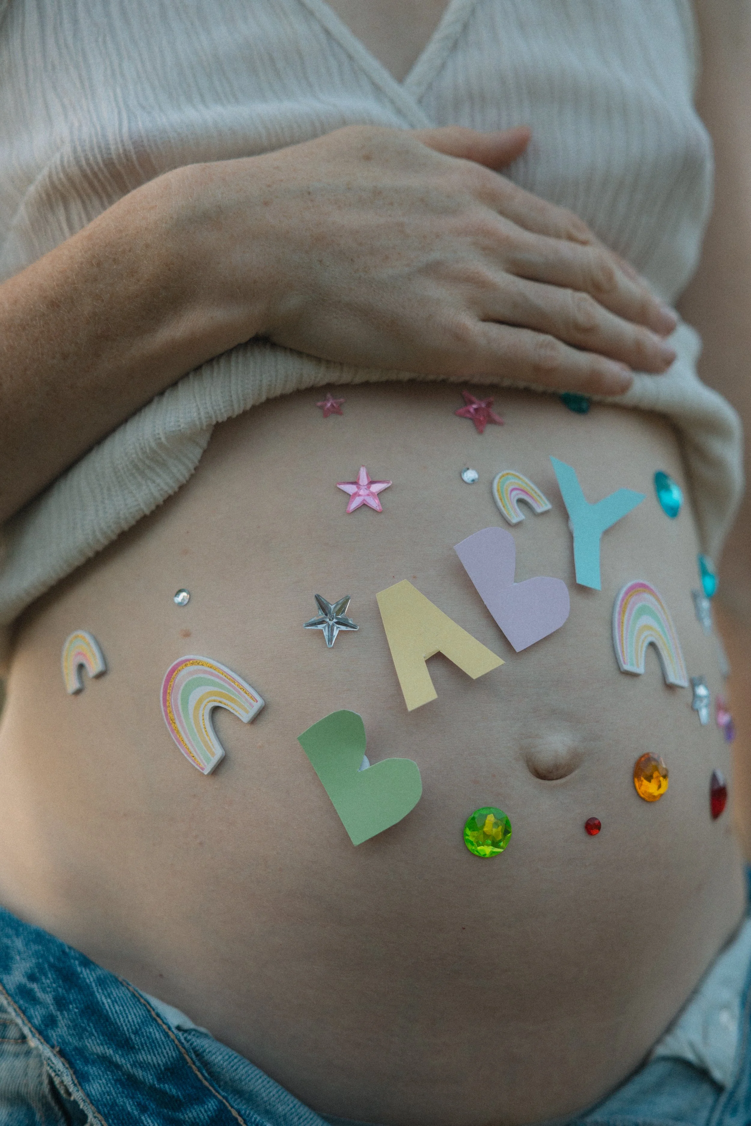 Decorated pregnant belly with colorful letter and rainbow stickers, surrounded by stars and gemstones.