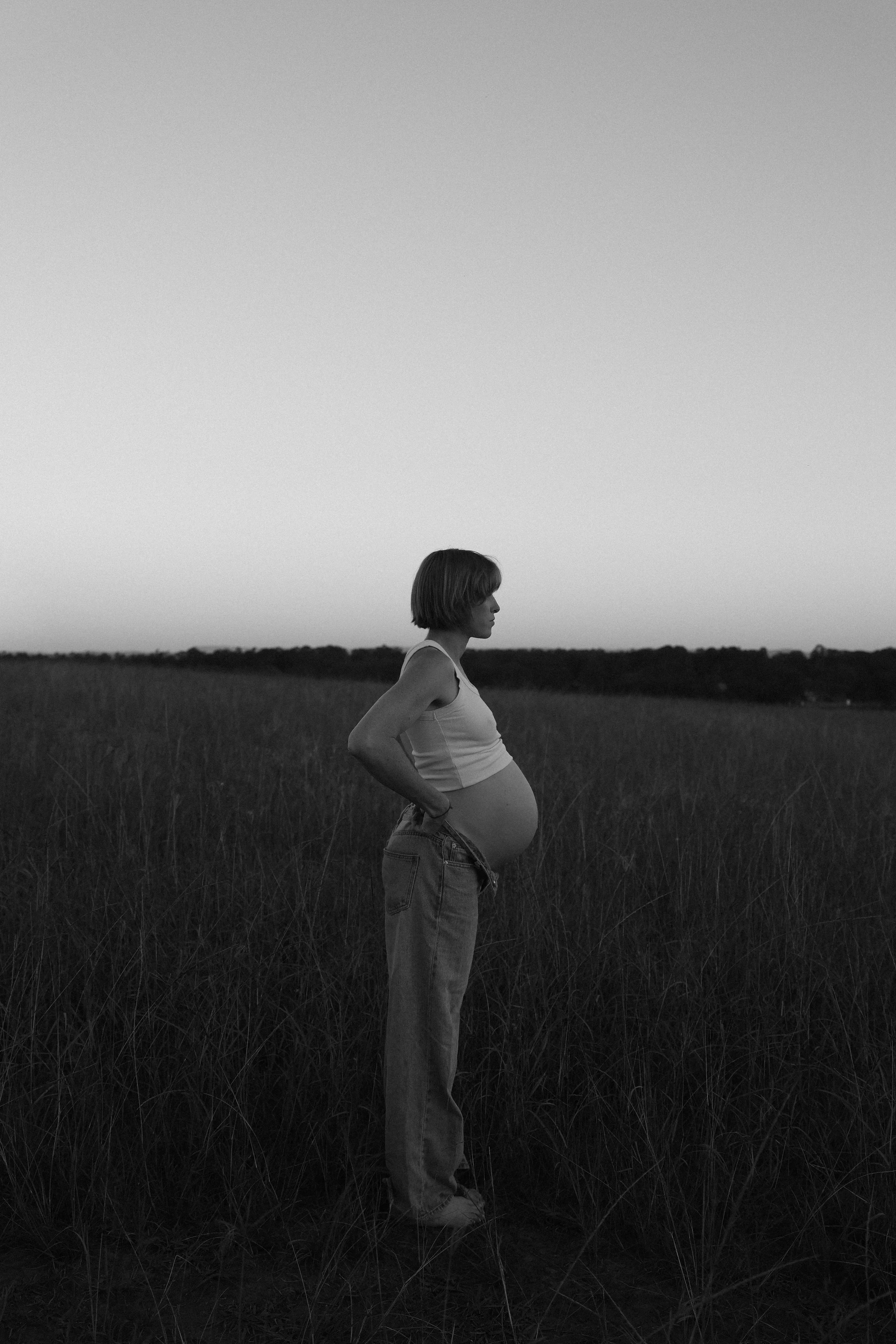 Black and white photo of a pregnant woman standing in a field with tall grass, facing to the right, with her hand on her hip, during dusk or night.