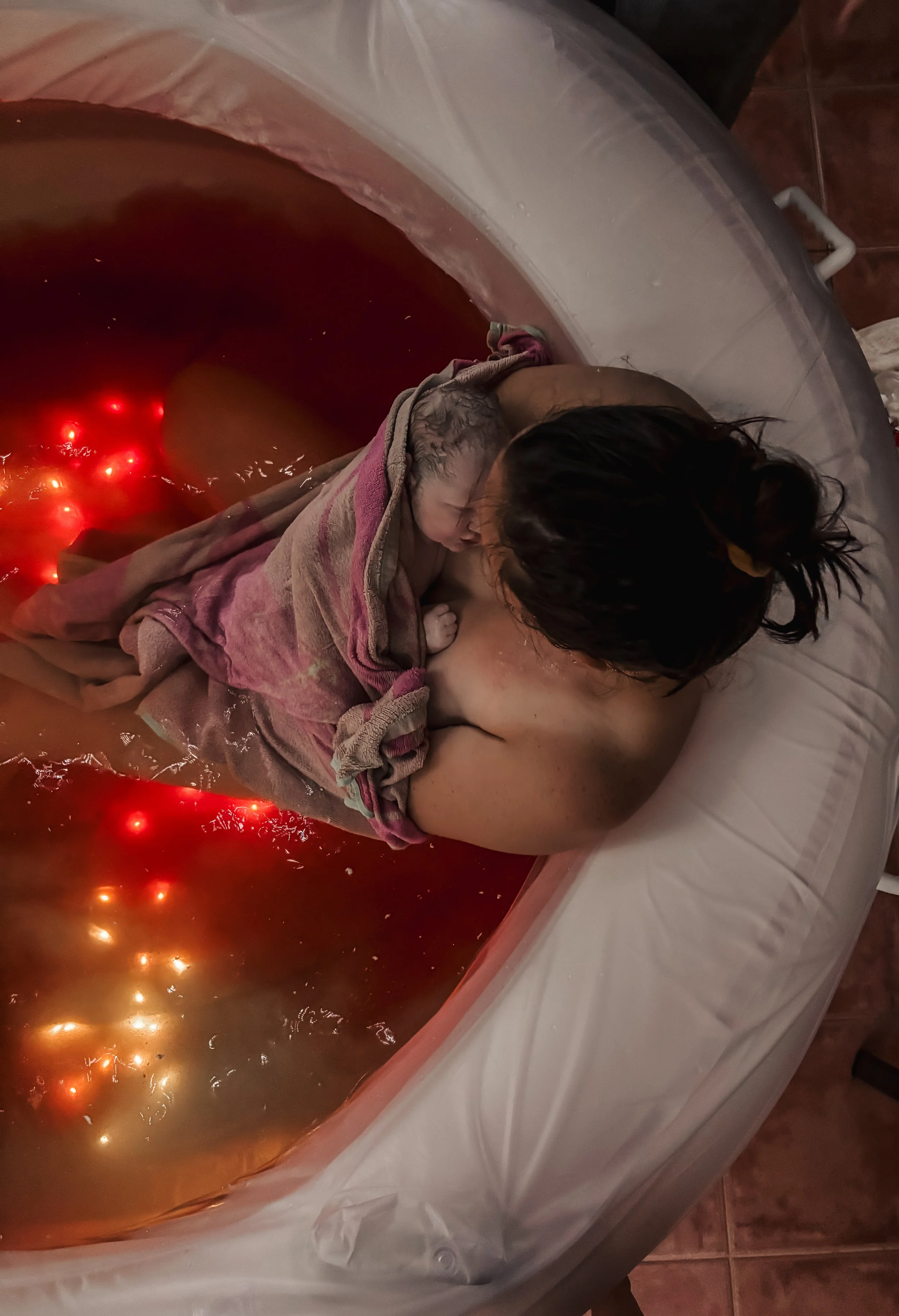 A mother giving birth to a baby in a birthing pool filled with water, illuminated by red lights and reflection, with the baby after delivery wrapped in a towel.