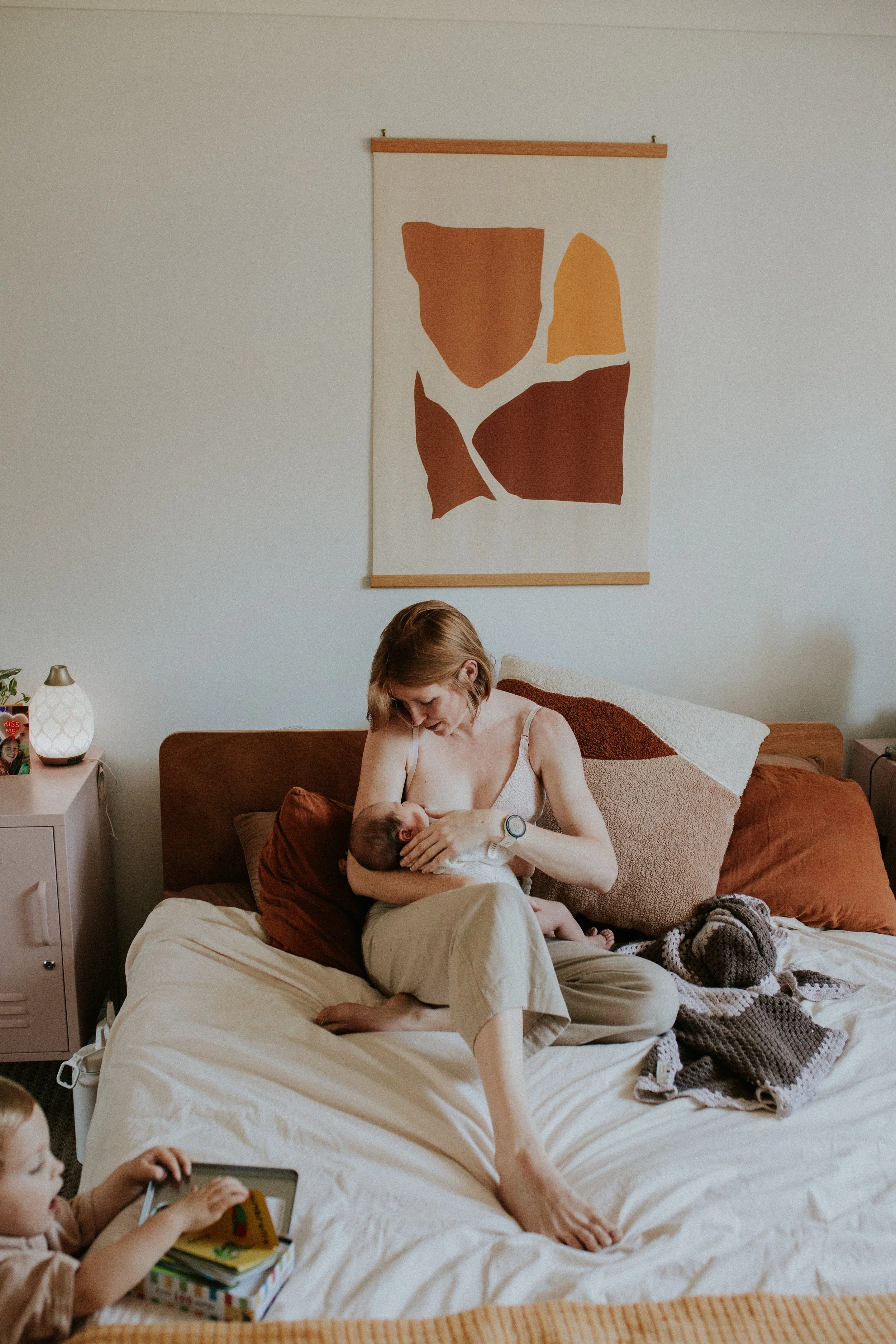 A woman sitting on a bed breastfeeding a baby, with a young child playing nearby.