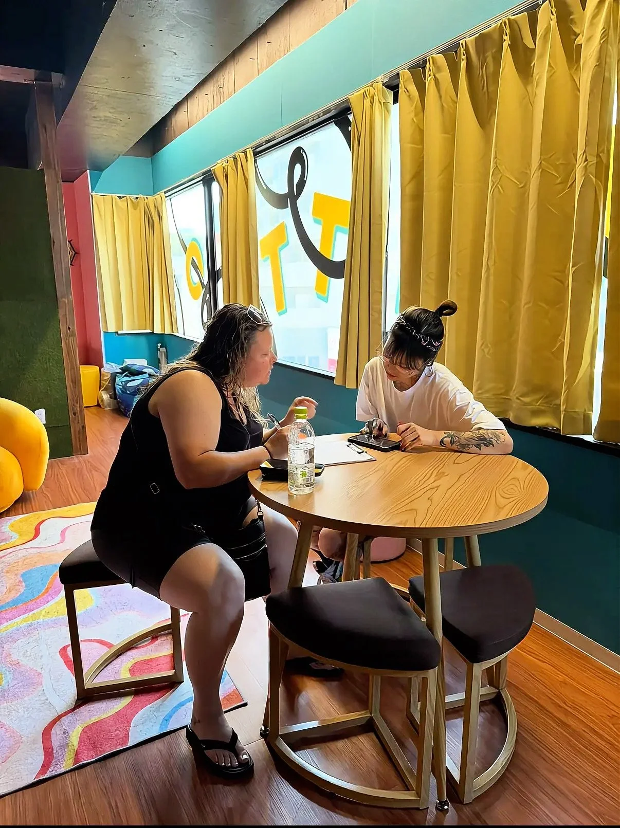 Two women sitting at a round wooden table in a colorful room One woman has light skin, dark curly hair, and is wearing a black dress and sandals. The other woman has light skin, dark hair in a bun, glasses, a white shirt, and tattoos on her arm. The room has yellow curtains, blue walls with a colorful message, and a multicolored rug on the wooden floor.