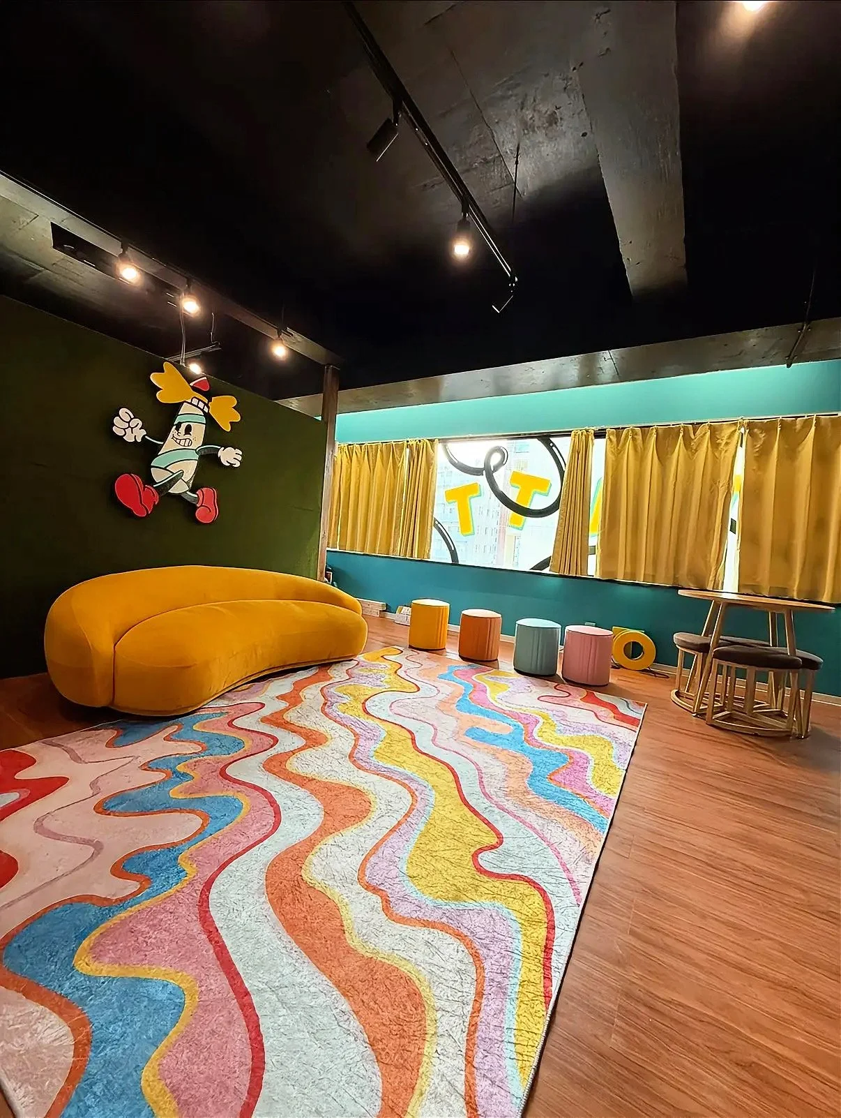 Colorful interior room with a multicolored swirled rug, a yellow sofa, small round stools in pastel colors, a wooden table with chairs, yellow curtains, a window with bright letters, and a cartoon character wall decoration.