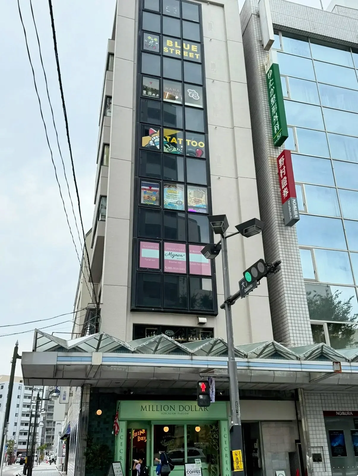 A tall building with multiple signs on the windows, including 'Blue Street' and 'Tattoo'. There is a green traffic light with a crosswalk signal and a green storefront labeled 'Million Dollar' at the entrance, with people walking underneath. There are overhead street wires and a clear sky.