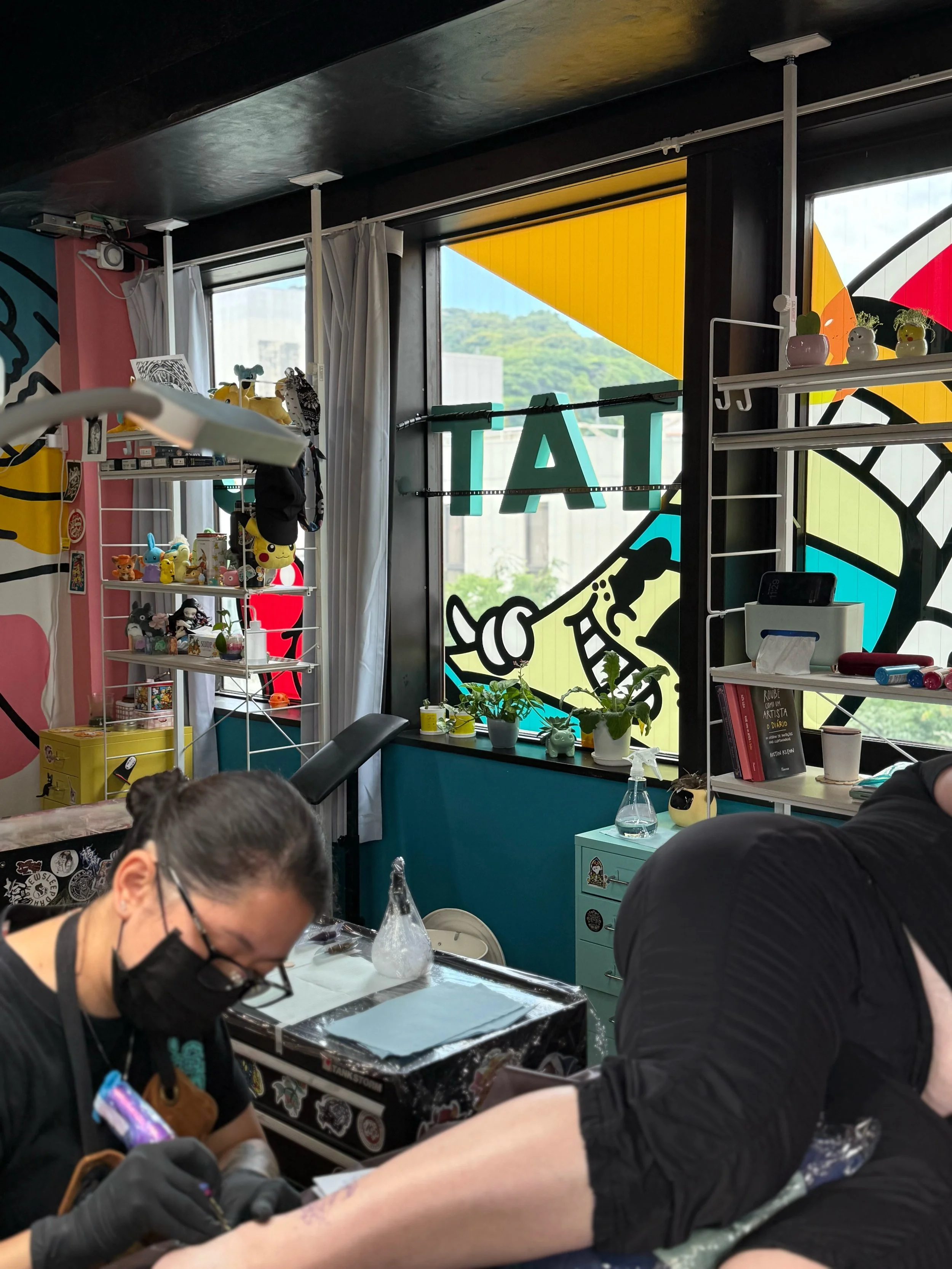 People tattooing on a person's arm inside a tattoo studio with colorful stained glass windows, shelves with small decorative items, and a woman wearing a face mask working on the tattoo.