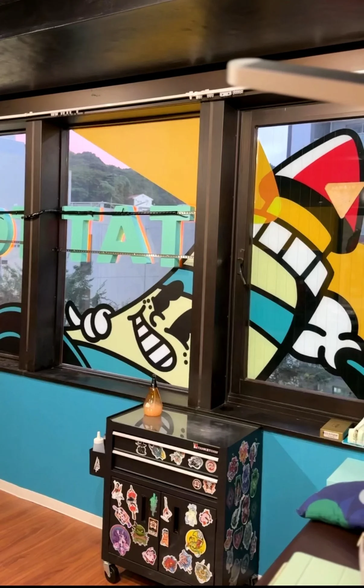 Decorative window with colorful cartoon-style artwork of a boat and a smiling person, inside a room with a blue wall, a black sticker organizer with various stickers, and a small table with a lamp.