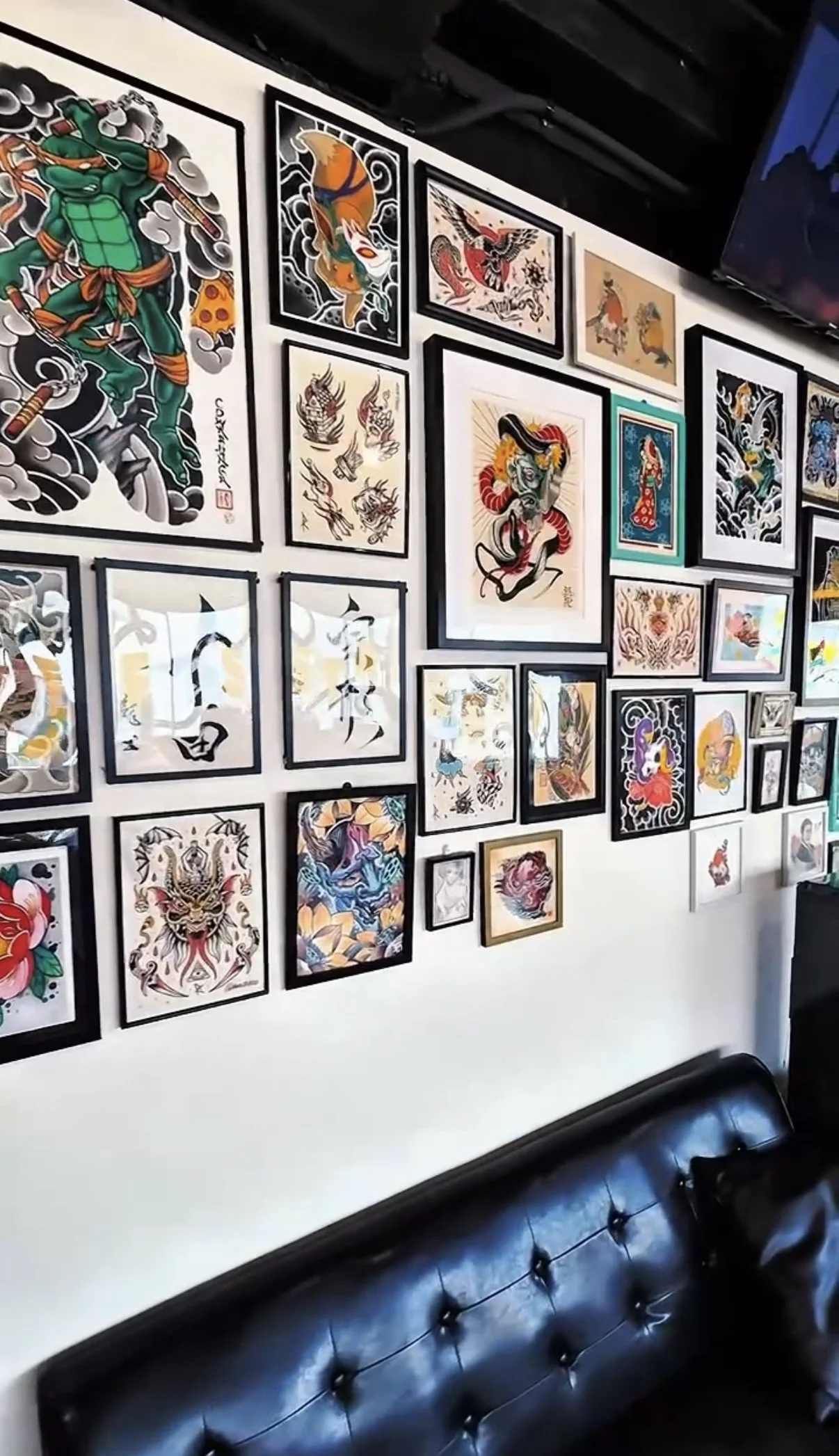 A wall decorated with multiple framed traditional and modern Japanese tattoo art, including dragons, koi fish, and mythical creatures, with a black leather tufted sofa in front of the wall.