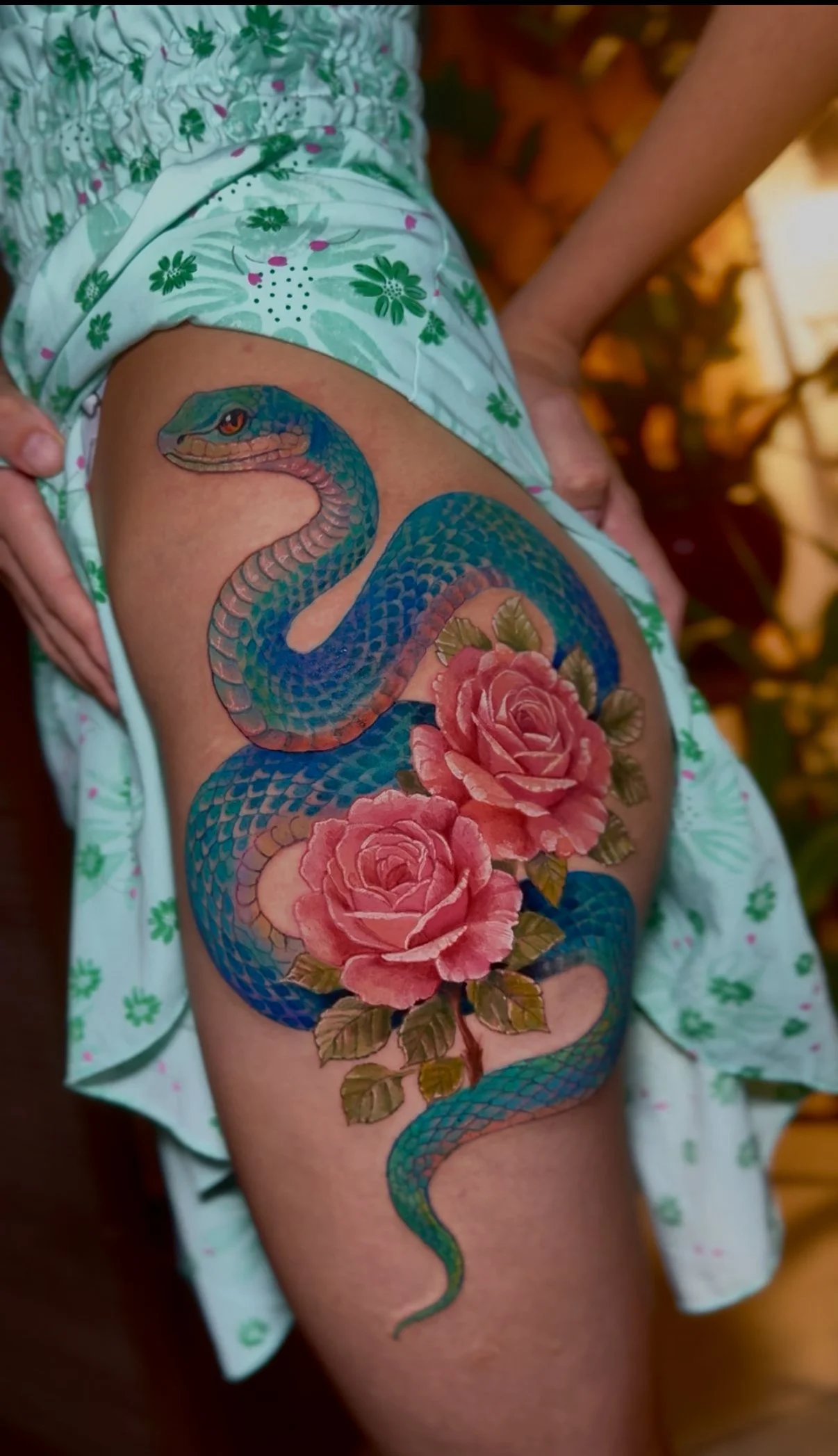 Colorful snake tattoo with roses on person's thigh, partially covered by floral shorts and a floral shirt.