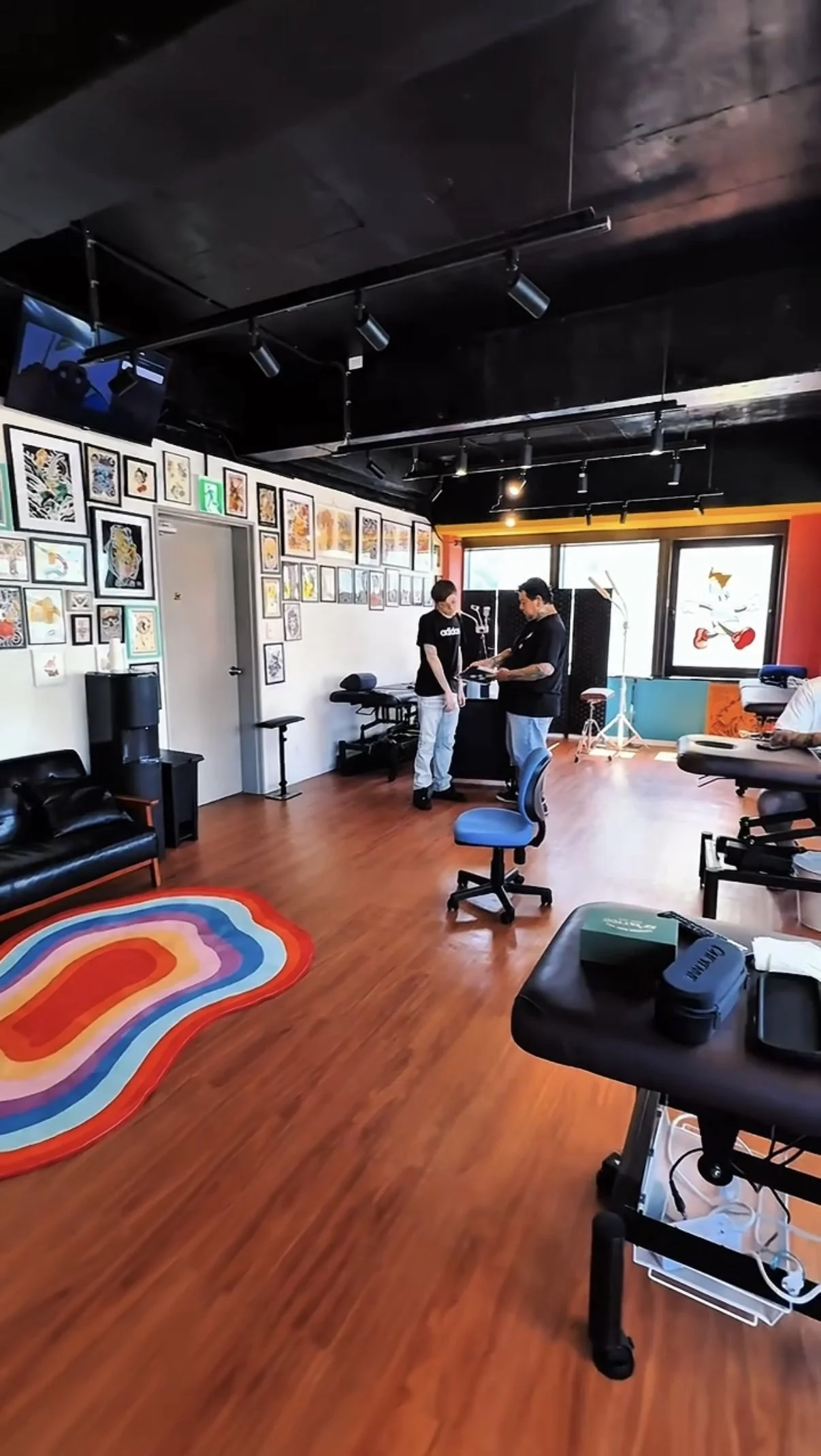 A colorful rainbow-shaped rug on a wooden floor in a tattoo studio. The studio has framed tattoo art on the wall, and two people are discussing something at a desk near a large window. There are chairs, a massage table, and tattoo equipment visible.