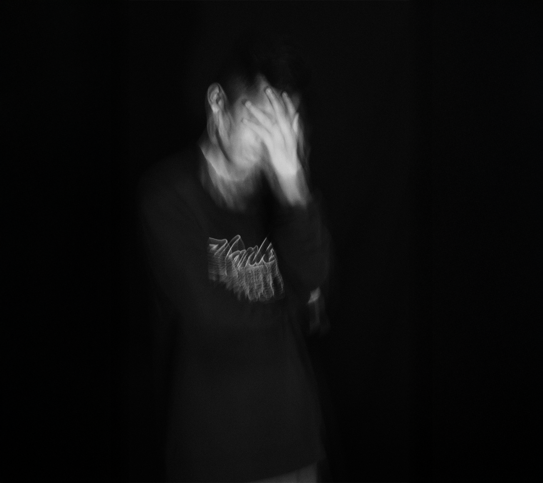 A person with short hair, wearing a dark sweatshirt, is covering their face with their hand in a dark setting, creating a blurred and emotional effect.