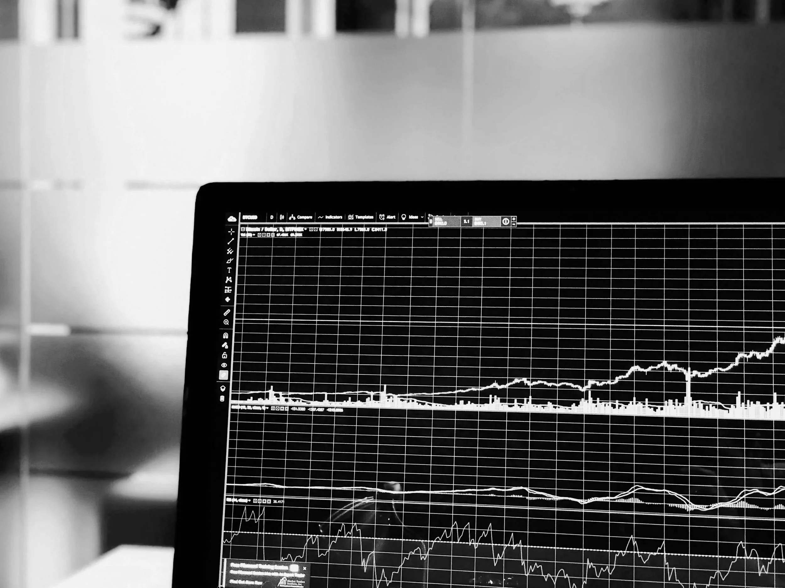 A computer screen displaying financial charts and data in black and white, with a background of blurred office furniture.