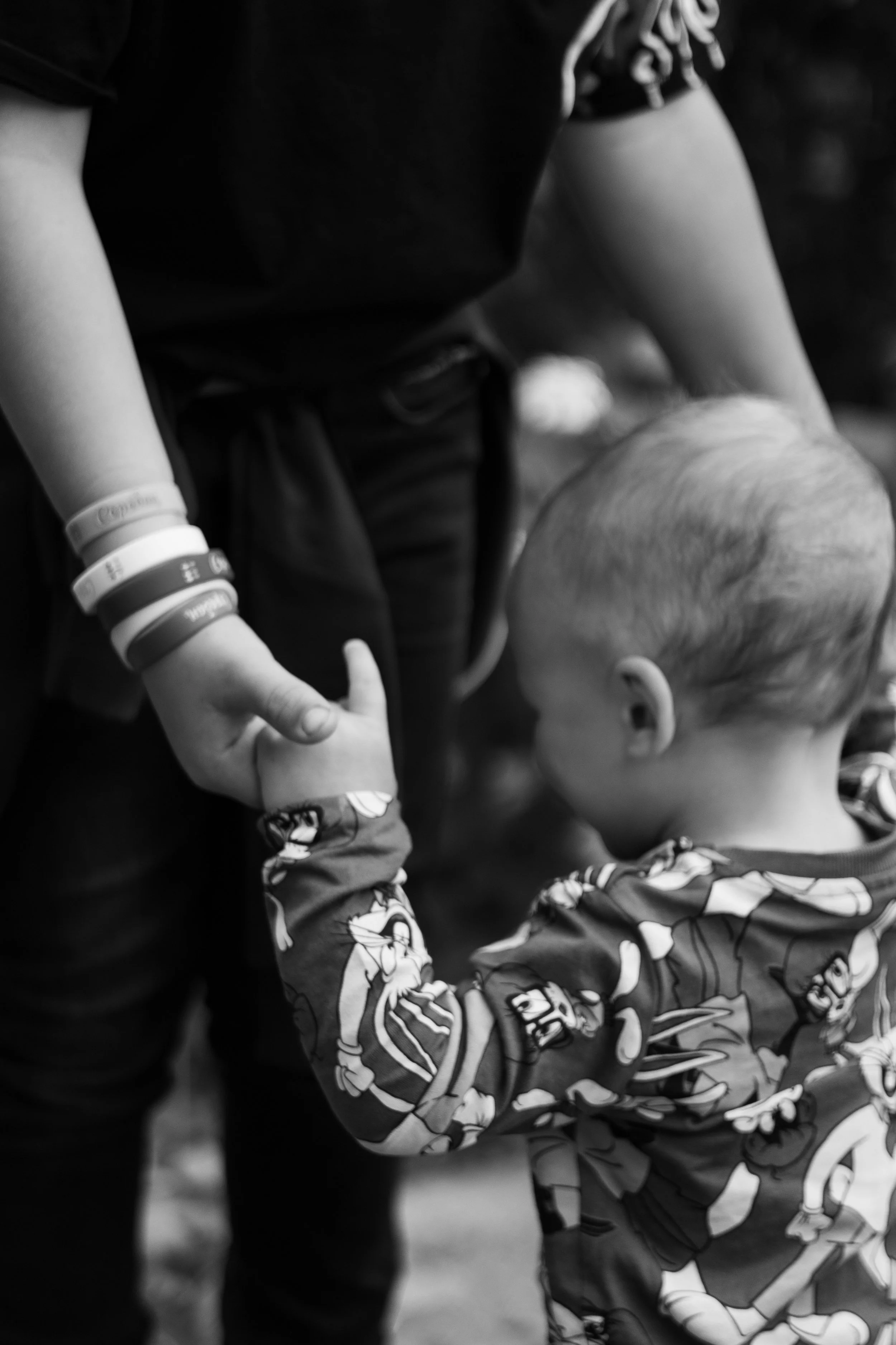 A young child with short hair, wearing a superhero-themed hoodie, holding hands with an adult, who is wearing several wristbands and a black outfit.