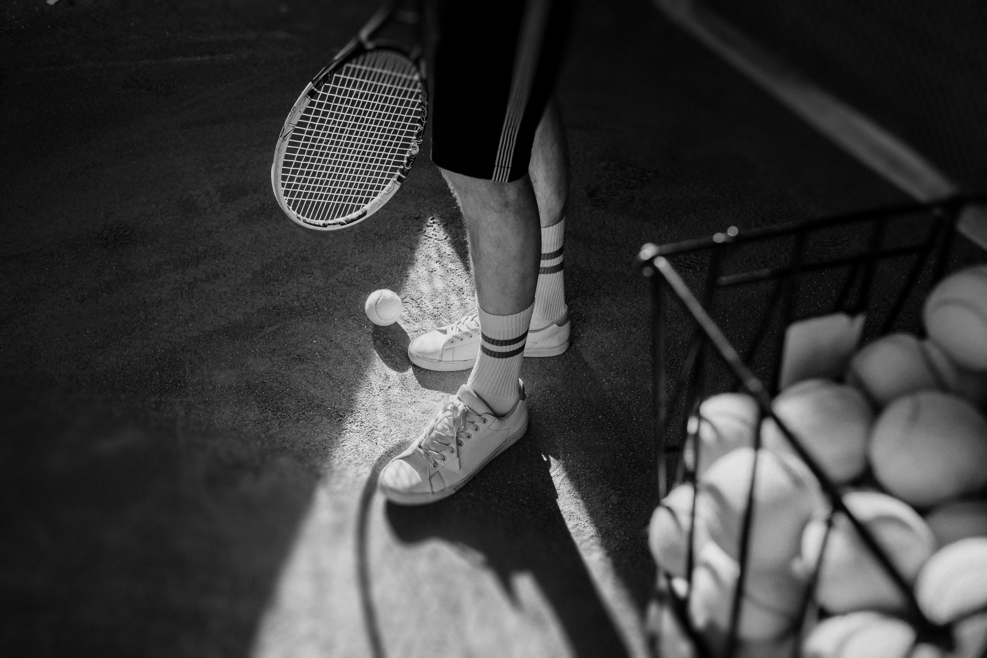 Close-up of a person standing on a tennis court, holding a tennis racket, with tennis balls in a container nearby.