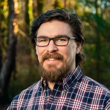 A man with glasses and facial hair smiling outdoors with trees in the background.